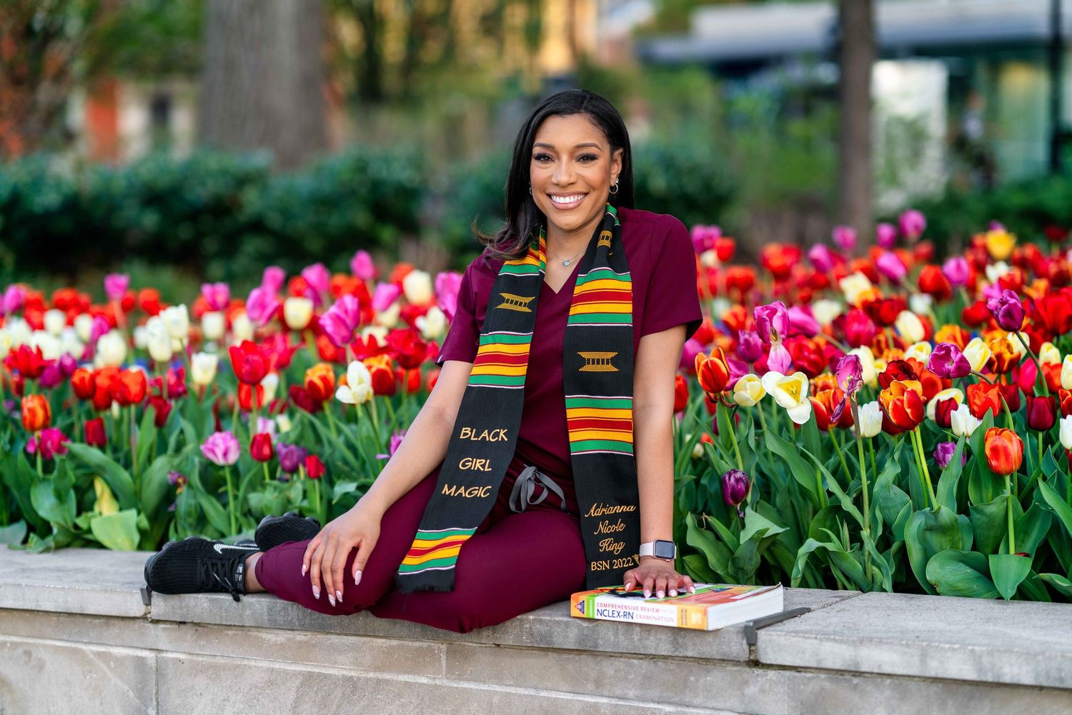 A female college graduate in a nursing stole and graduation cap, sitting cross-legged in front of a vibrant flower bed.