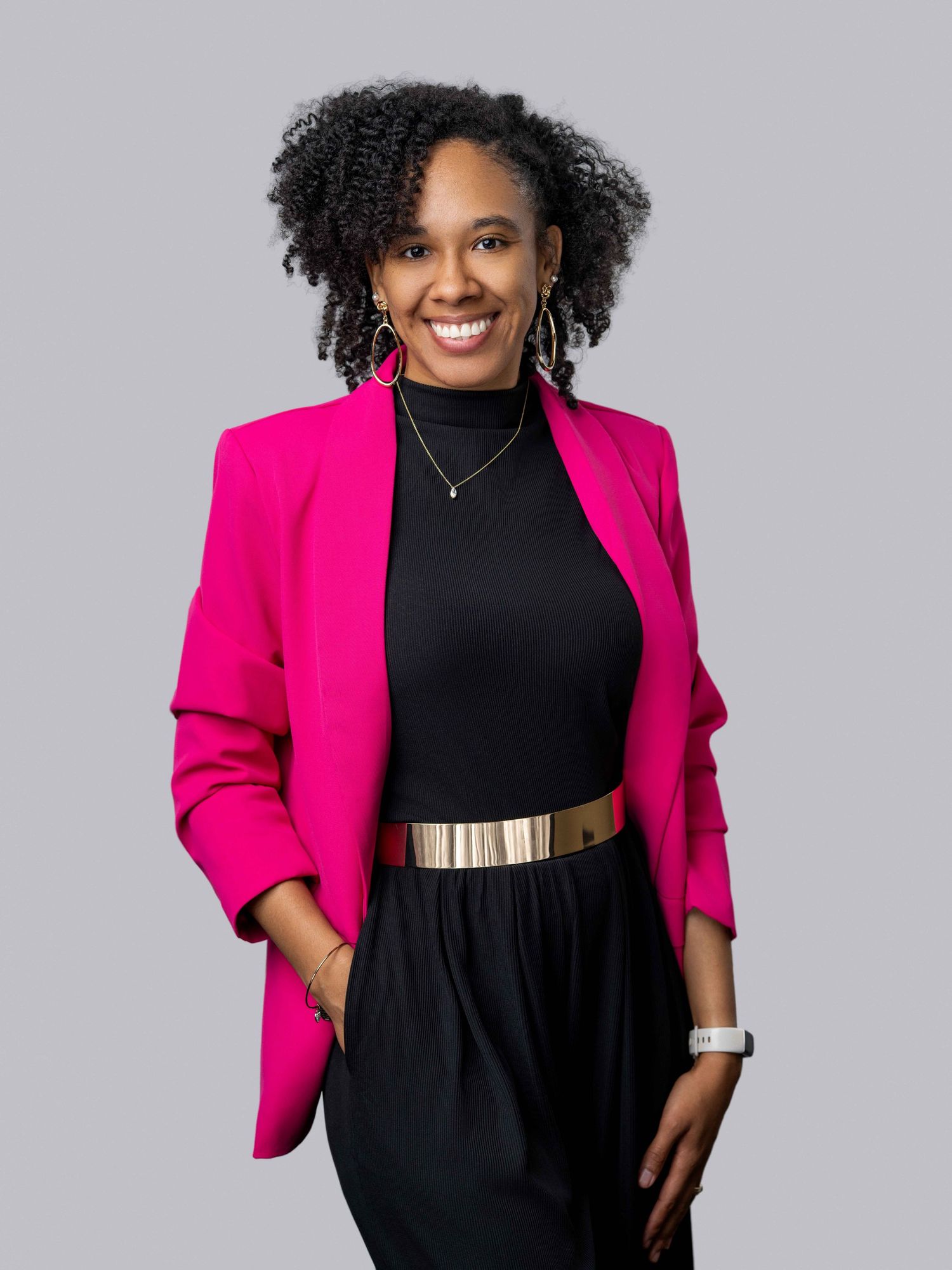 Studio portrait of an African American woman with curly hair, wearing a bright pink blazer and black trousers.