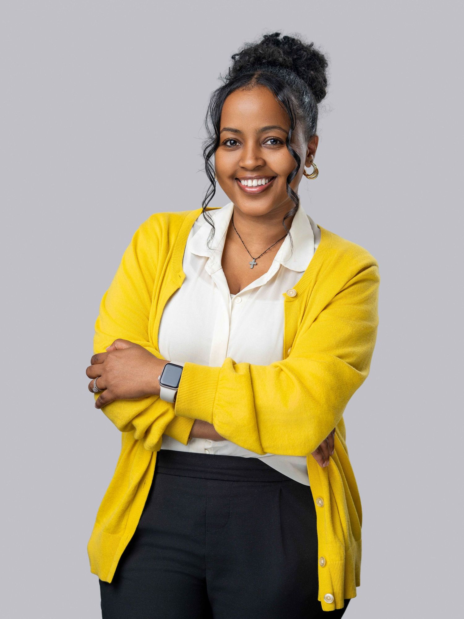 Close-up professional headshot of a smiling woman in a yellow blazer, posed with arms crossed against a gray studio backdrop.