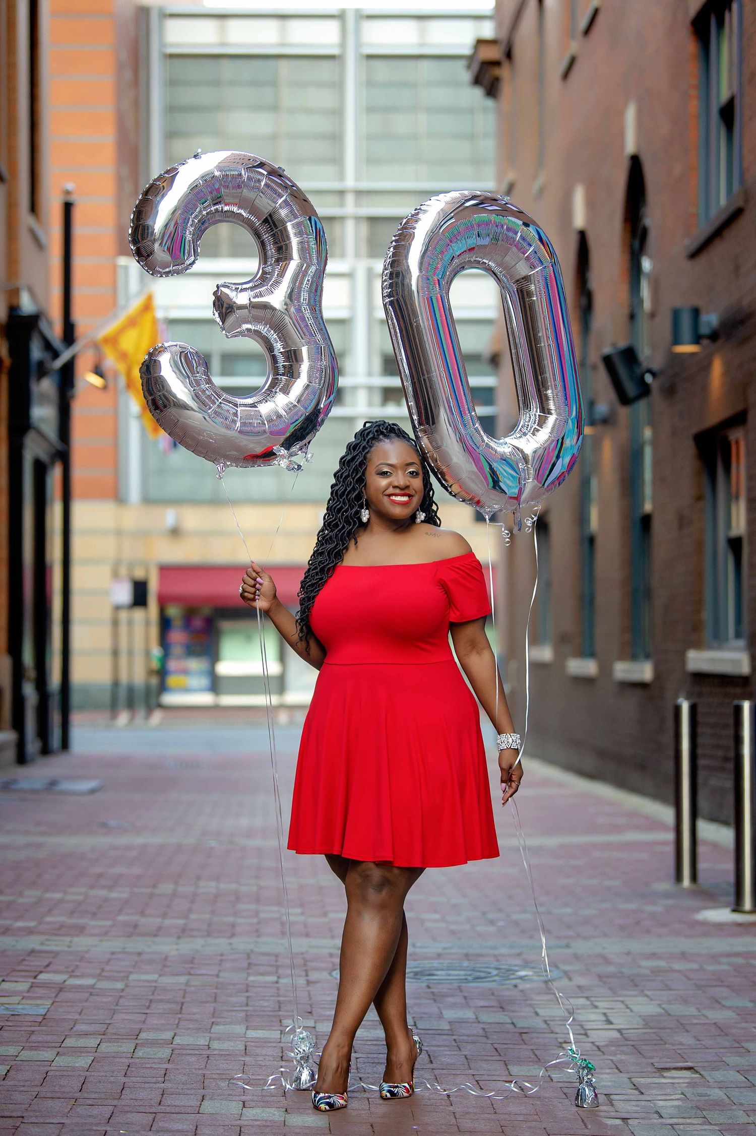 African american female standing in alley way, holding balloons for her 30th birthday in a red dress.