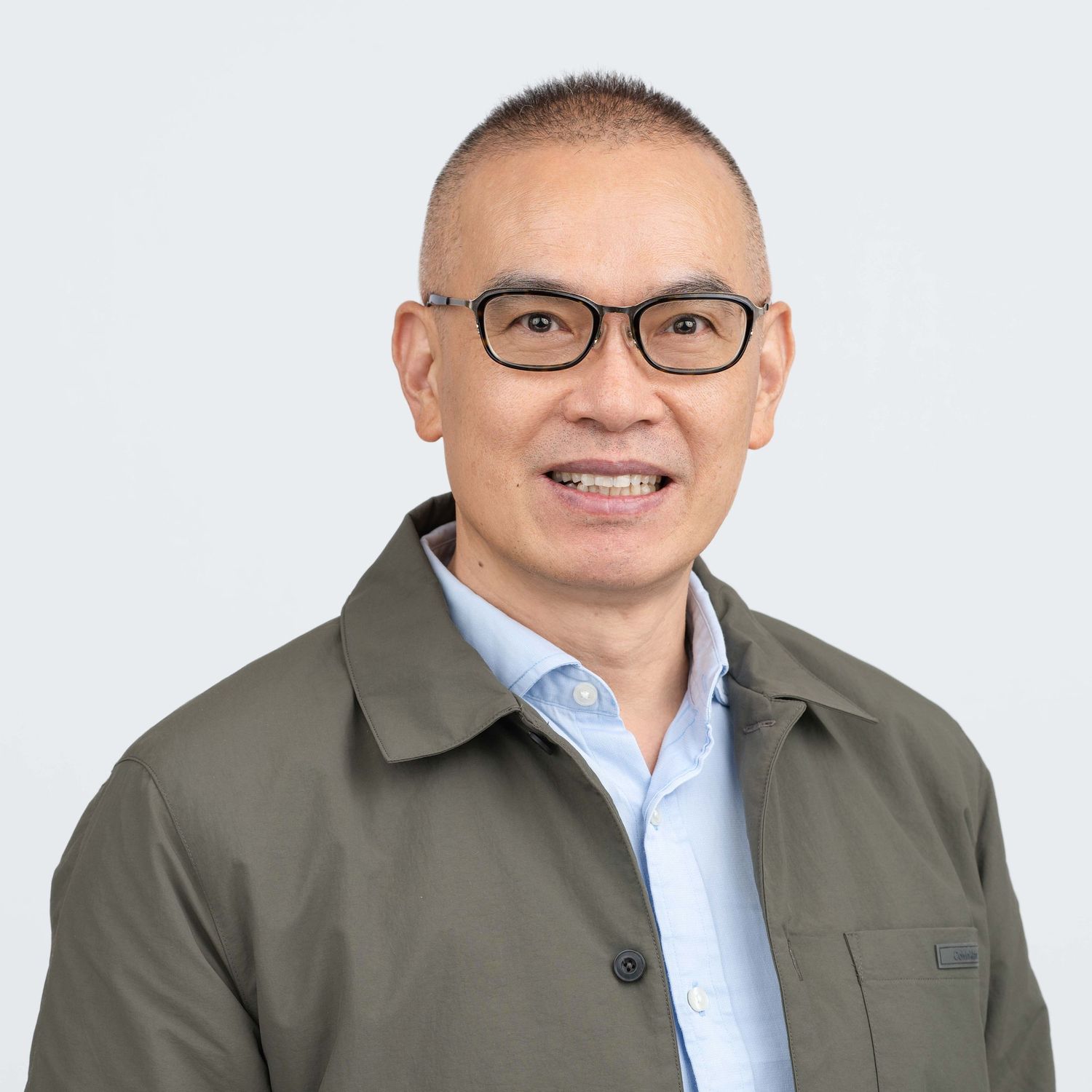 Professional headshot of a man with glasses, wearing an olive green jacket over a light-colored shirt.