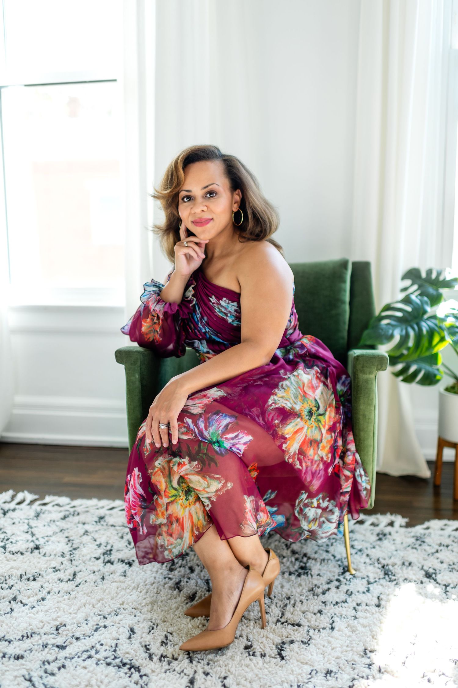 Elegant portrait of a woman in a floral dress sitting on a white chair in a bright, sunlit room.