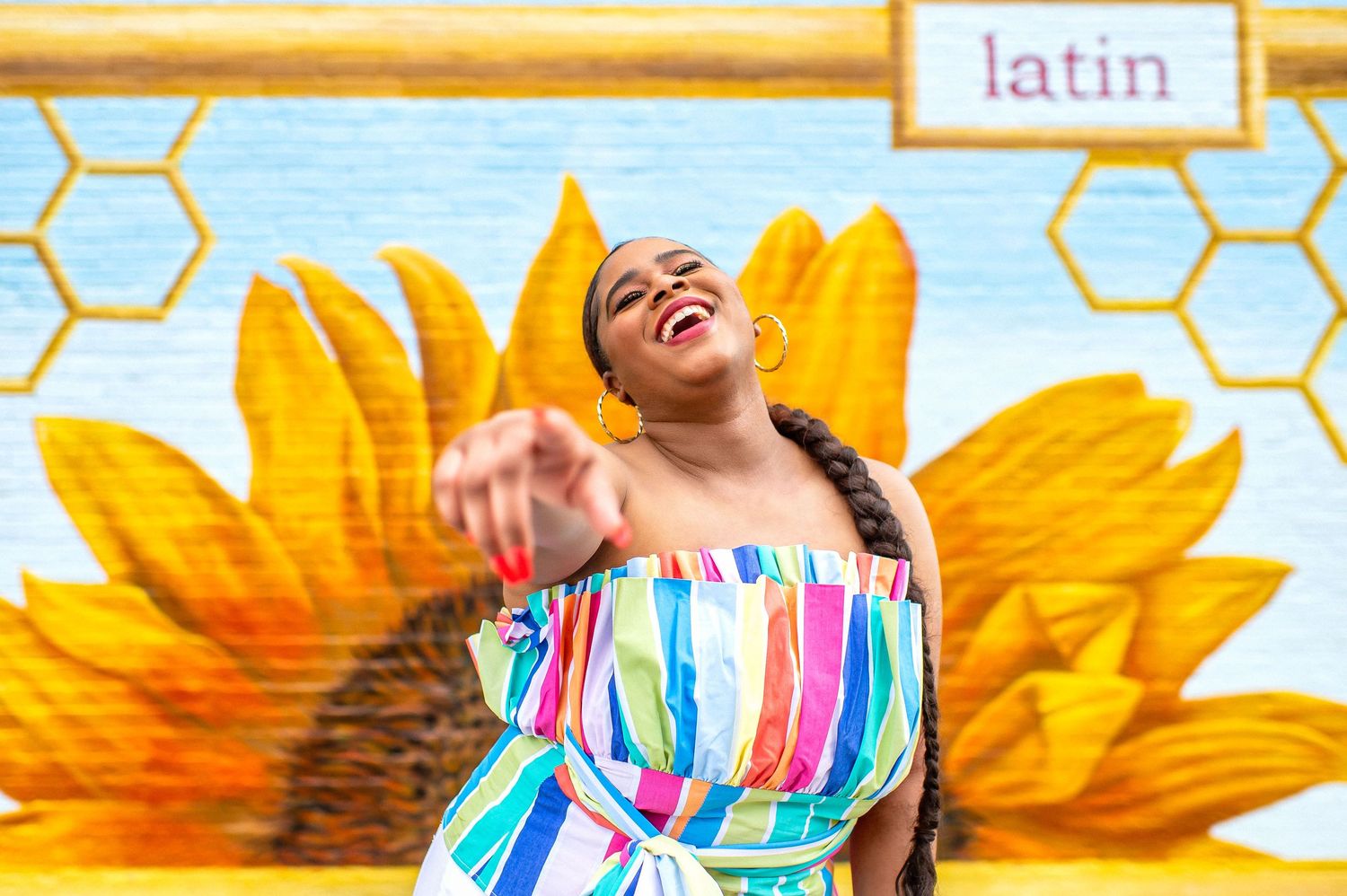A smiling person in a colorful striped dress points at the camera, posing before a sunflower mural with a honeycomb pattern and 