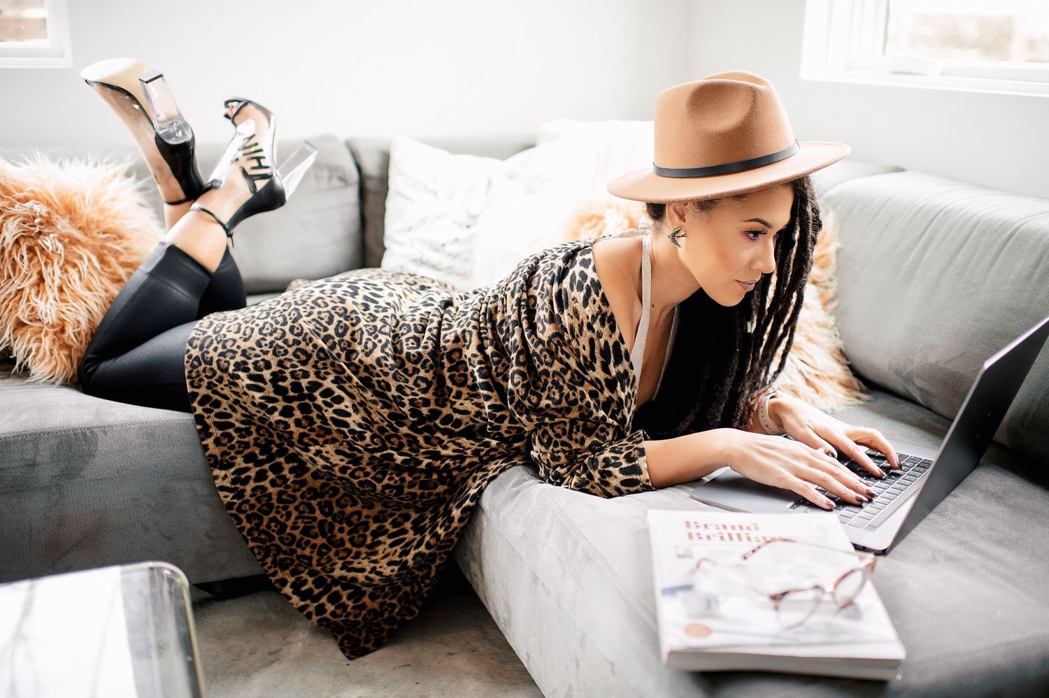 African american female wearing a hat, laying on a sofa working on a laptop with legs crossed.
