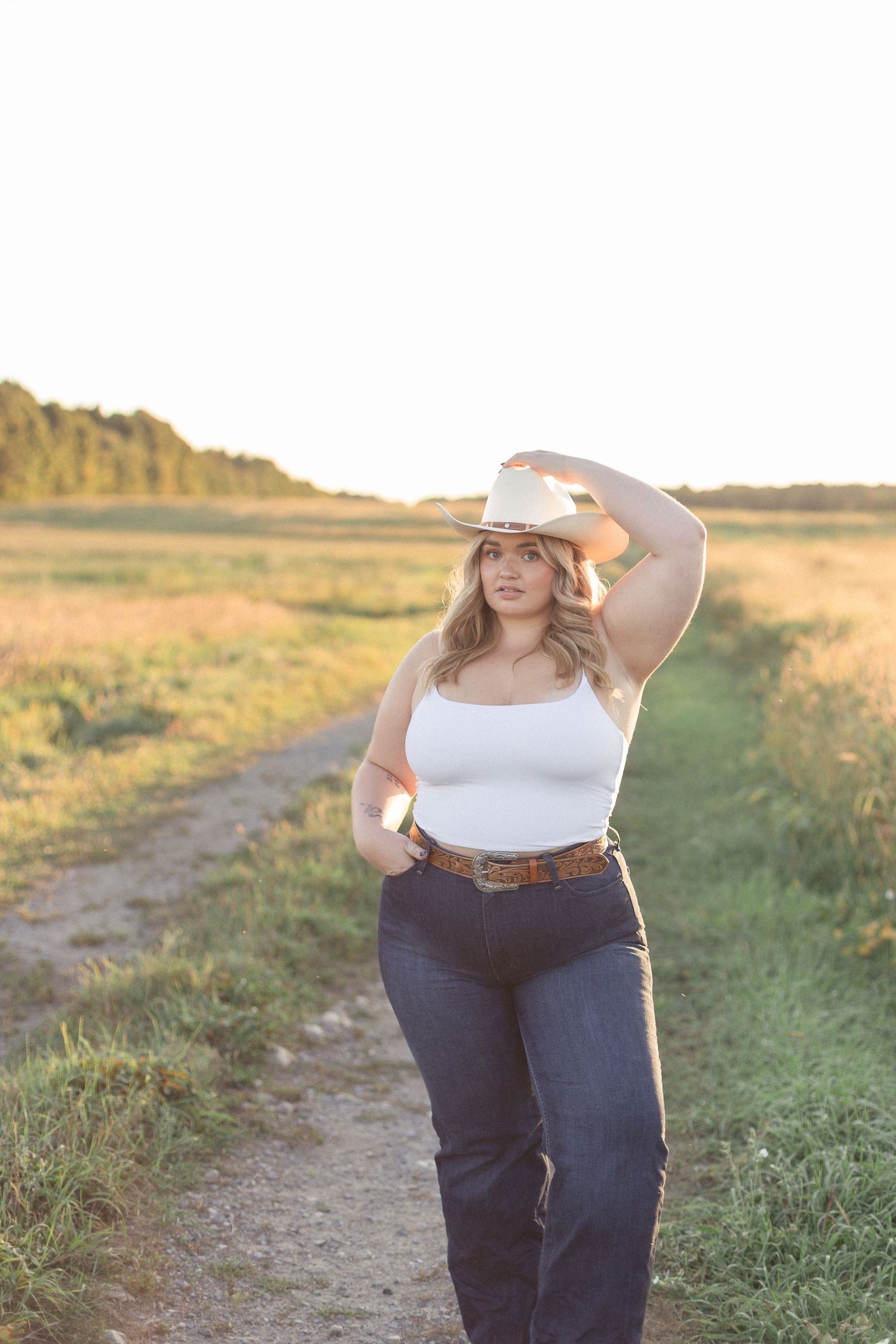 Femme debout dans les champs au coucher du soleil, tenant son chapeau de cowboy.
