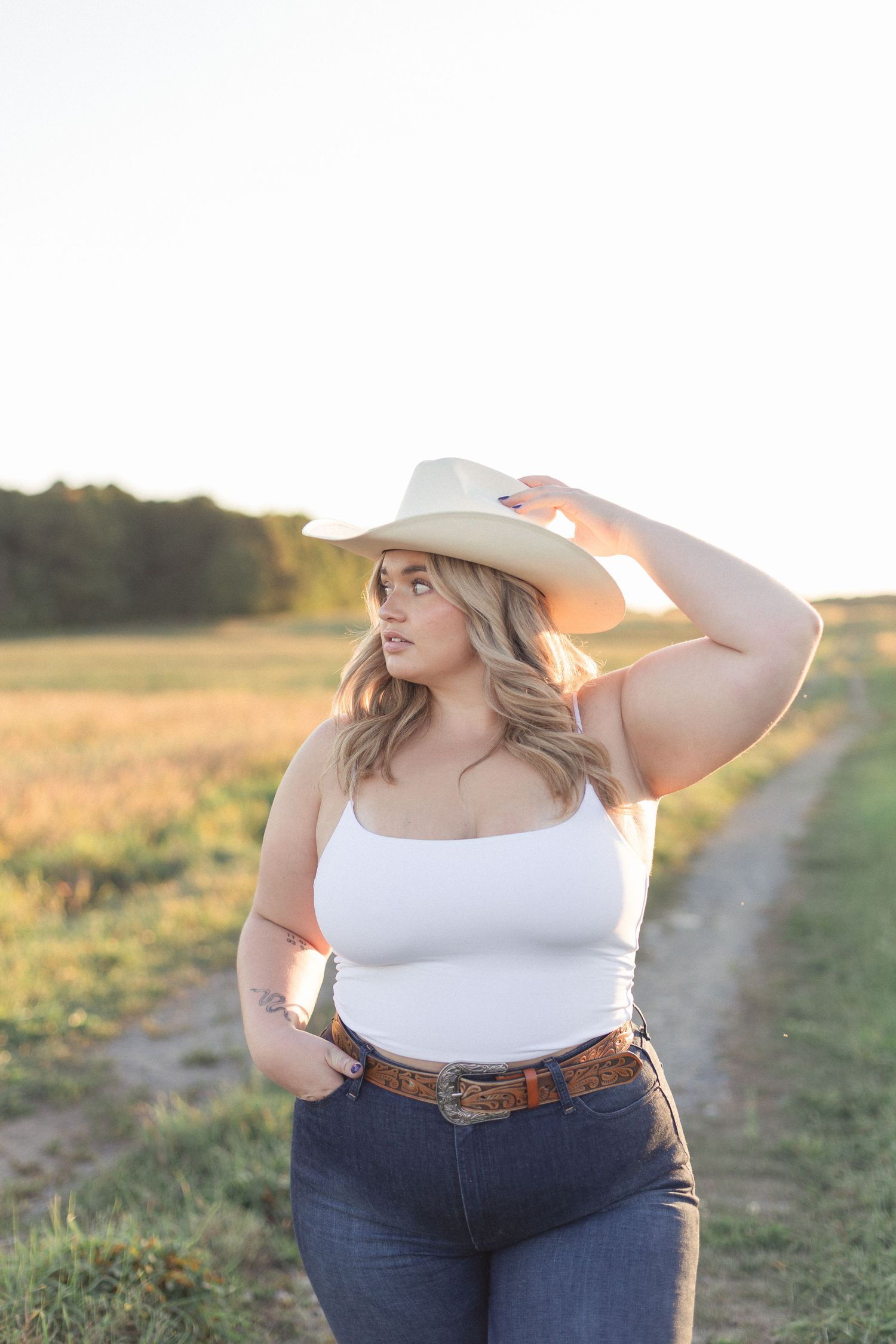 Femme dans un champ en lumière naturelle, la main sur son chapeau, regard tourné vers l'horizon.