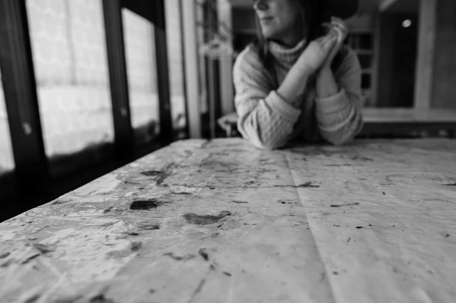 Photographie, en noir et blanc dramatique, d'une femme assise à une table remplie de taches de peinture.