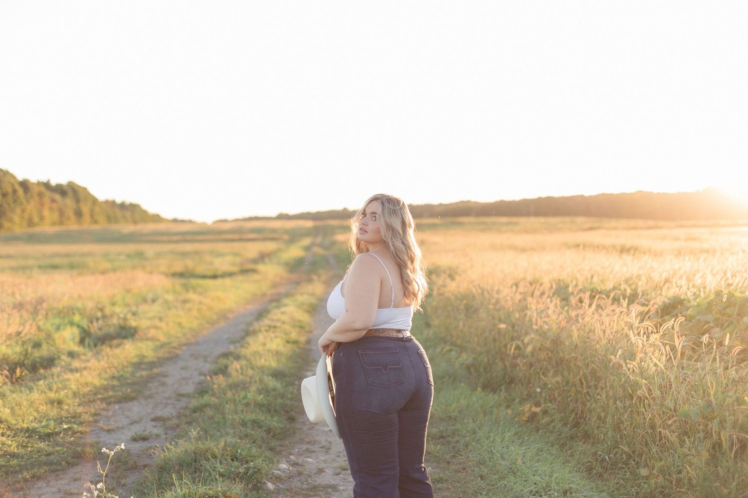 Femme debout sur un chemin dans les champs en automne, éclairée par la lumière douce du coucher du soleil.