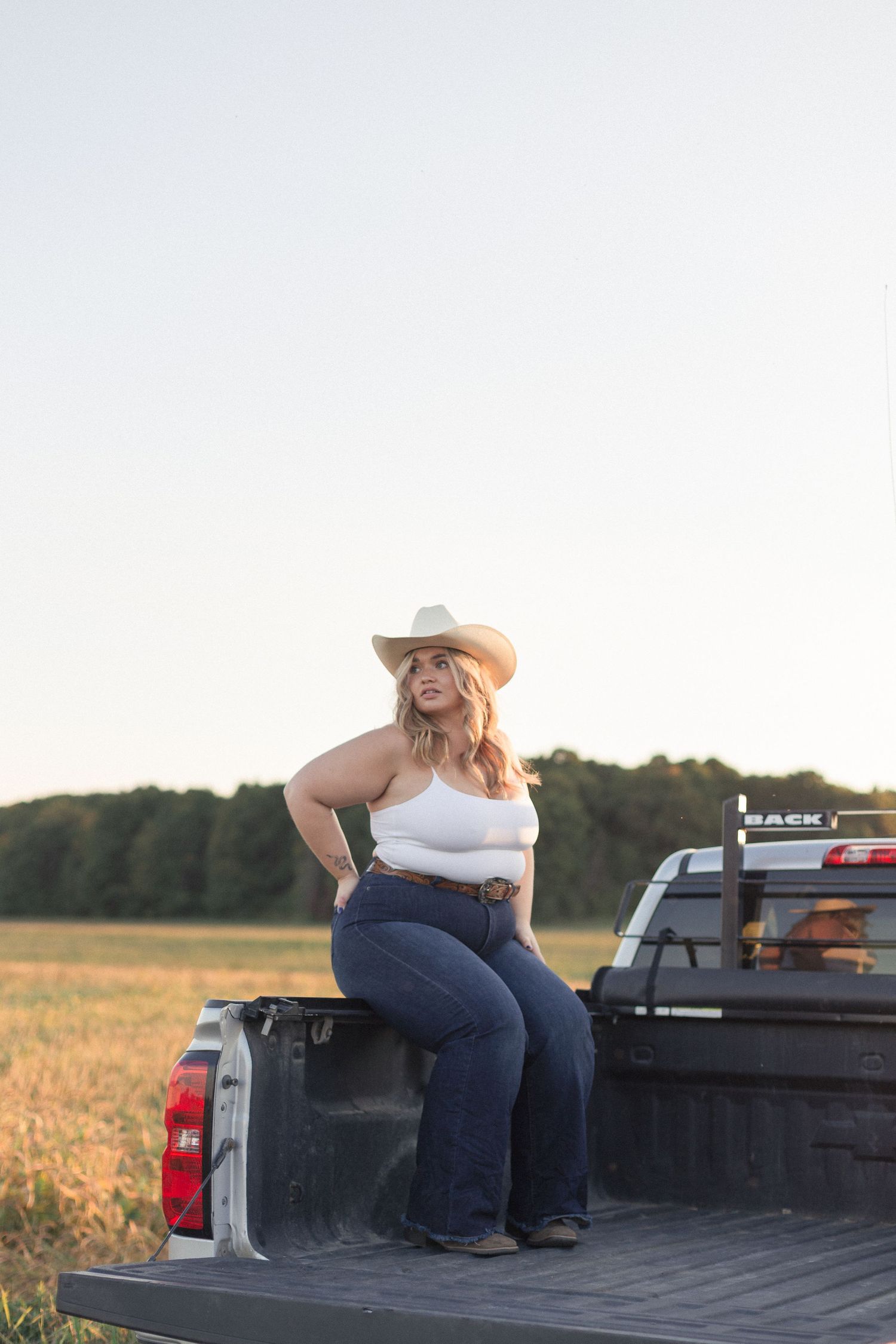 Portrait d'une femme assise dans la boîte d'un camion, portant un chapeau de cowboy et des jeans, en lumière naturelle.