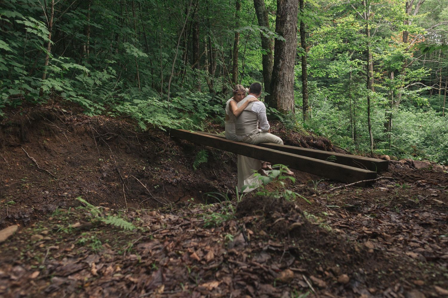 la mariée enlace son époux, assis sur un tronc d'arbre, au milieu de la forêt.