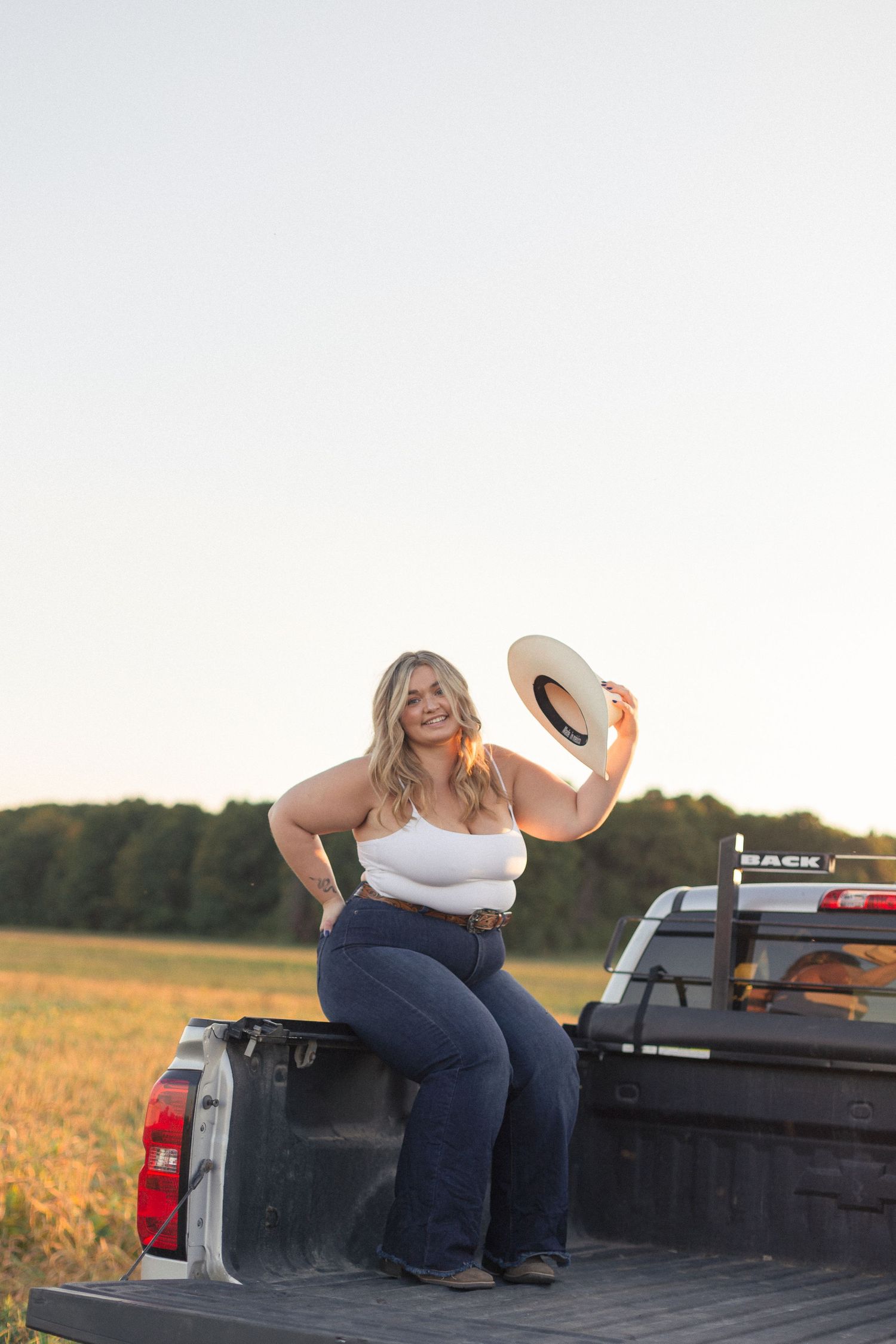 Une jeune femme en jeans tenant son chapeau de cowboy, assise dans la boîte d'un pick up, séance photo lifestyle.