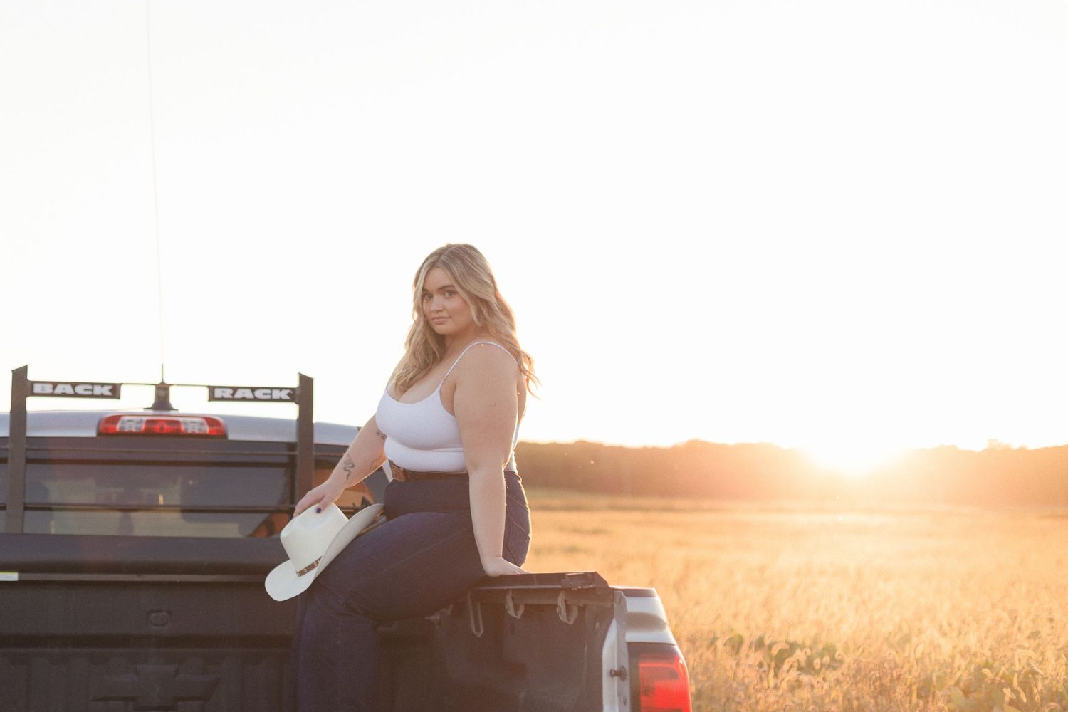 Femme assise dans la boîte d'un camion, qui tient son chapeau de cowboy, avec en arrière-plan, le soleil qui se couche à l'horizon.