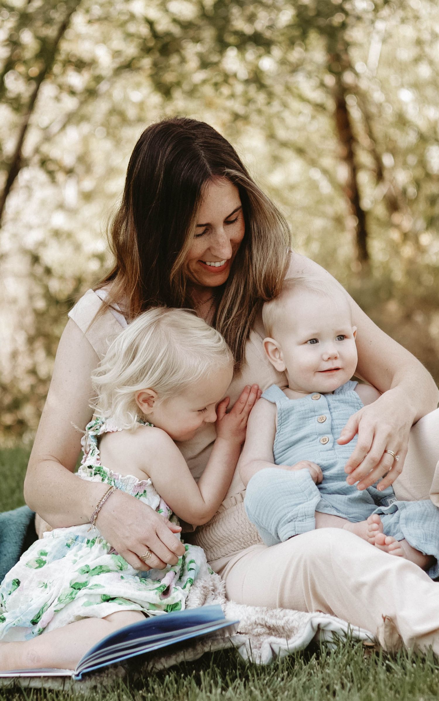 Mother and children reading a book together while laying on blanket outdoors.