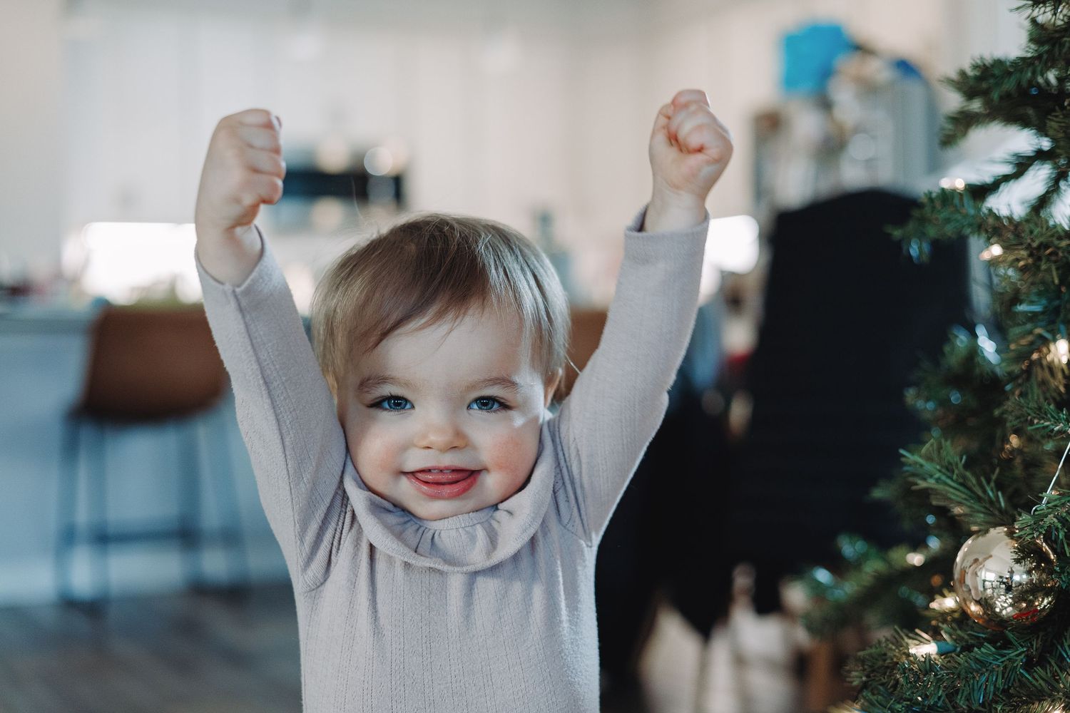 A young child raises their hands excitedly in front of a decorated Christmas tree while wearing a light gray sweater.