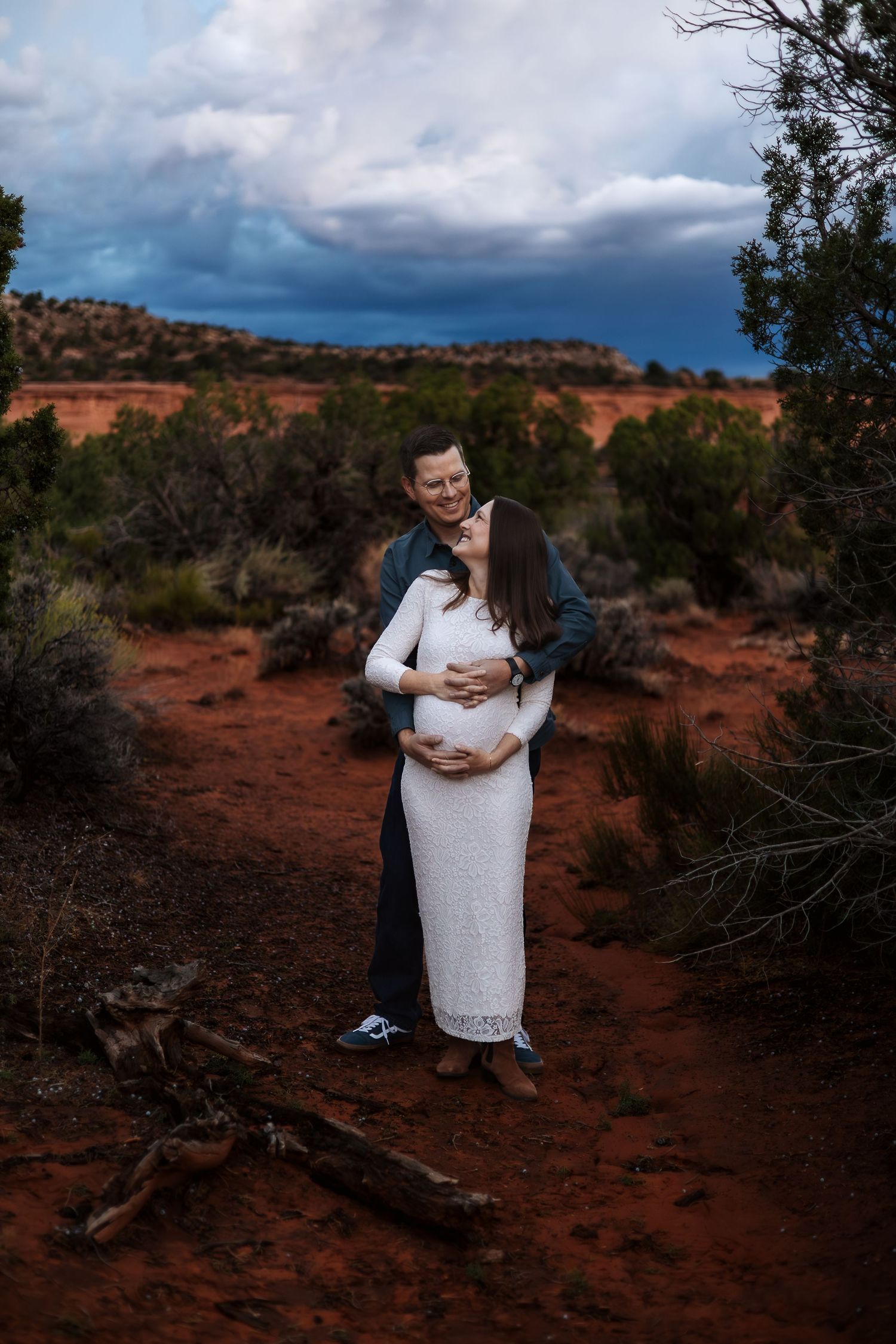 A couple embraces in a romantic moment against the backdrop of red desert soil and dramatic stormy skies in Utah.