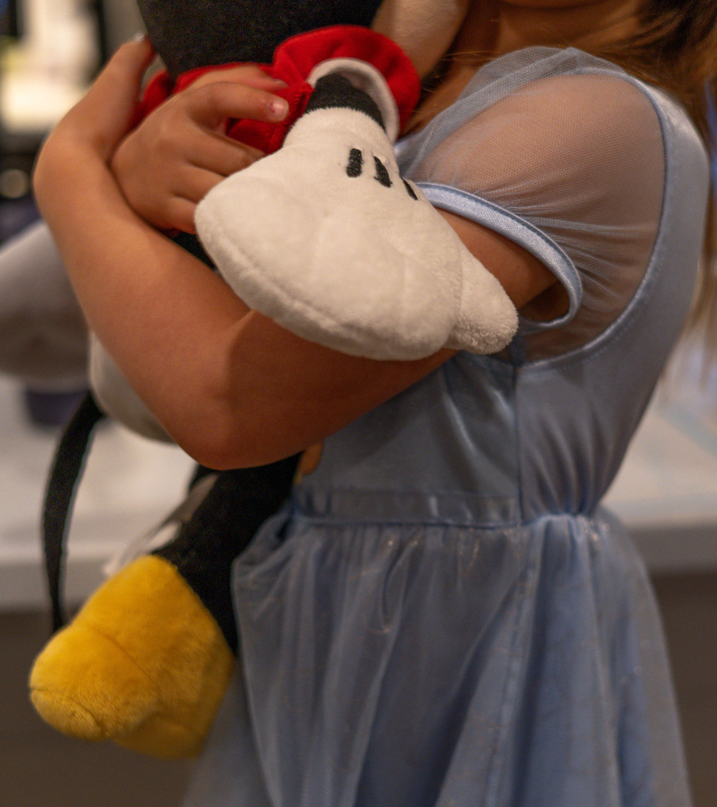 A person in a light blue dress holds a plush Mickey Mouse toy while standing indoors.
