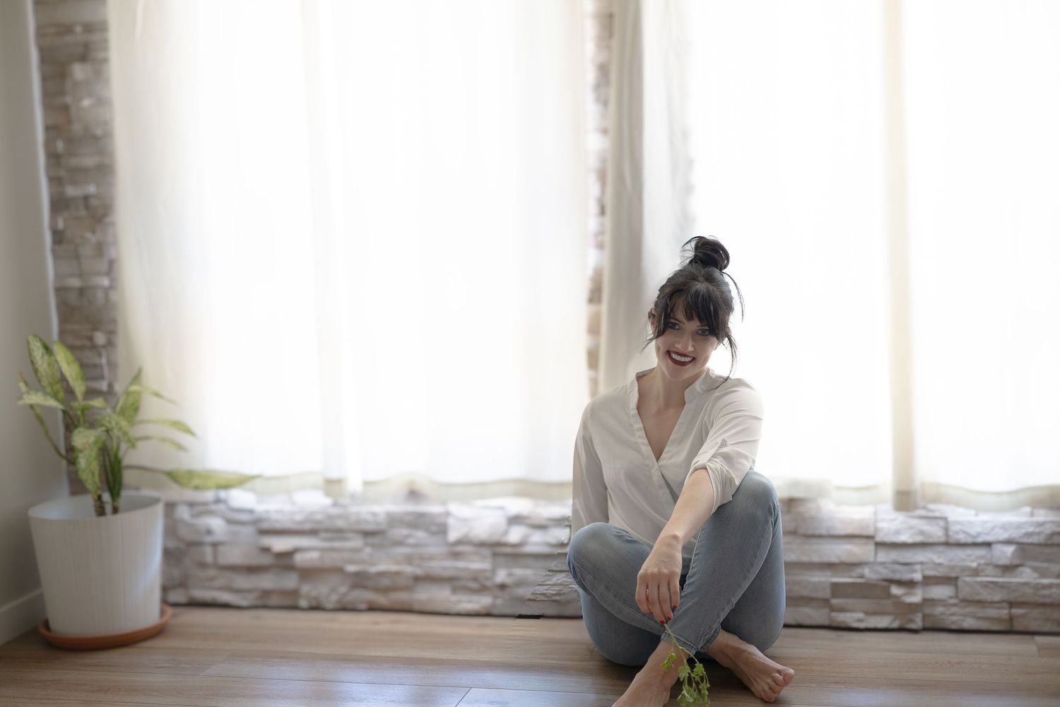 Window light illuminates figure in gray cardigan sitting beside potted plants in serene minimalist space.