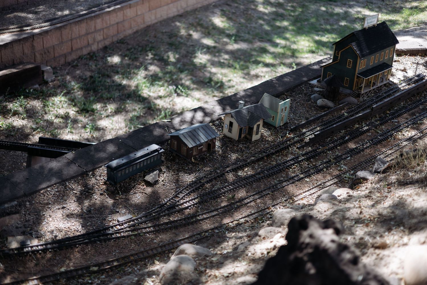 Miniature train track curves through a shaded garden setting with dappled sunlight filtering through the trees.