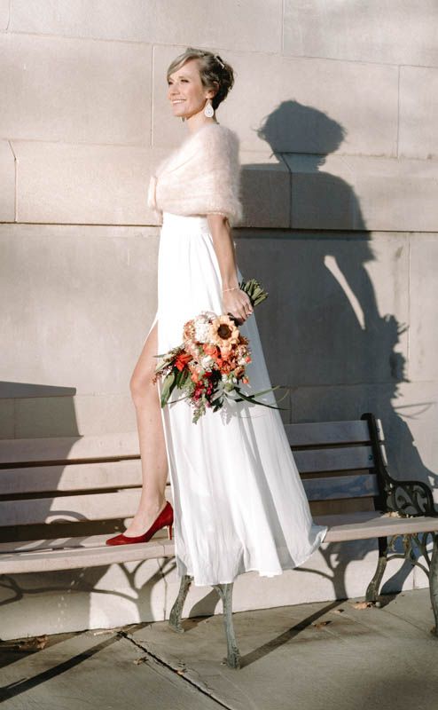 A bride in a white dress and red heels casts shadows on a light stone wall while holding a bouquet.