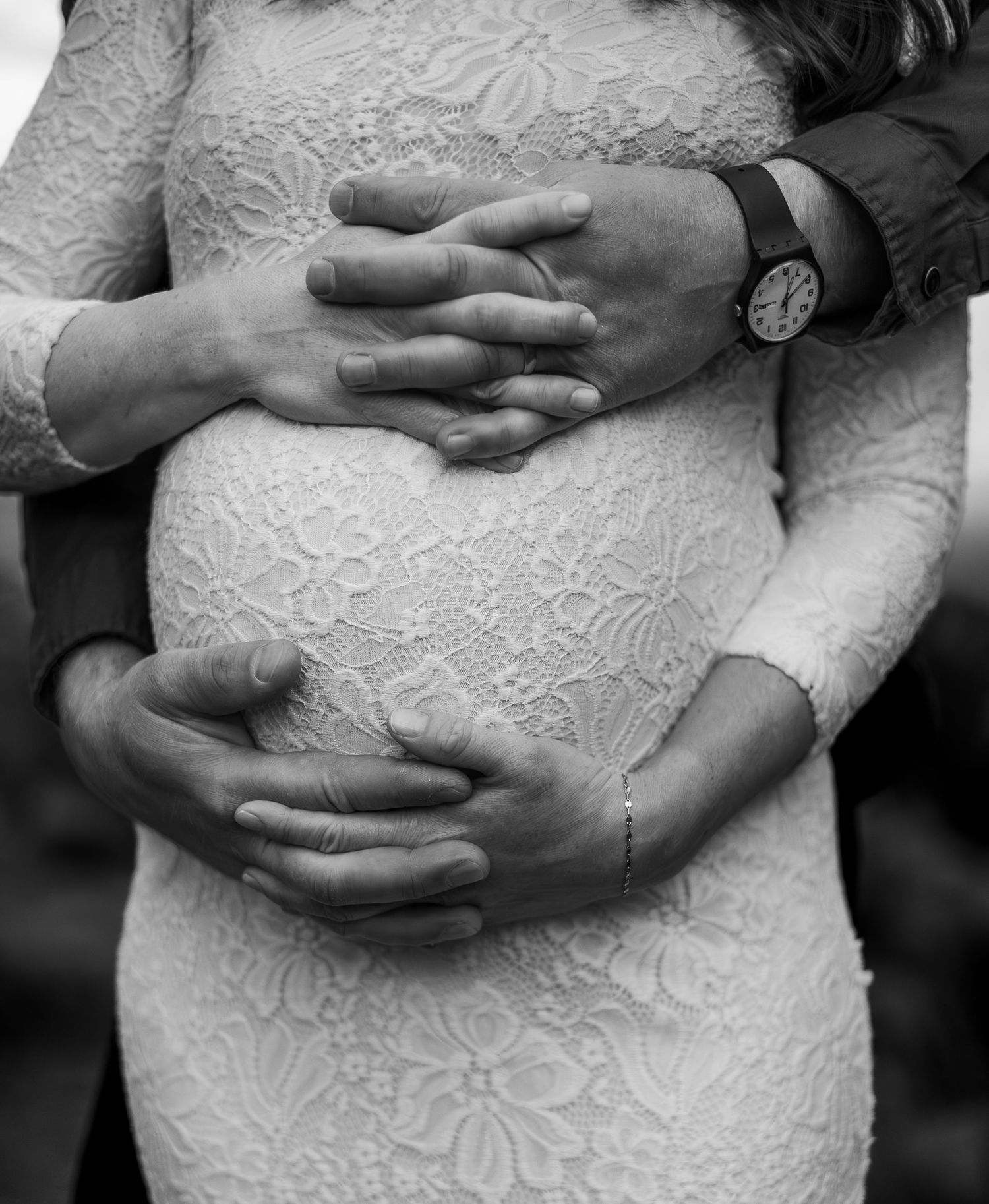 Intimate black and white photo showing hands tenderly cradling a pregnant belly in a lace wedding dress.
