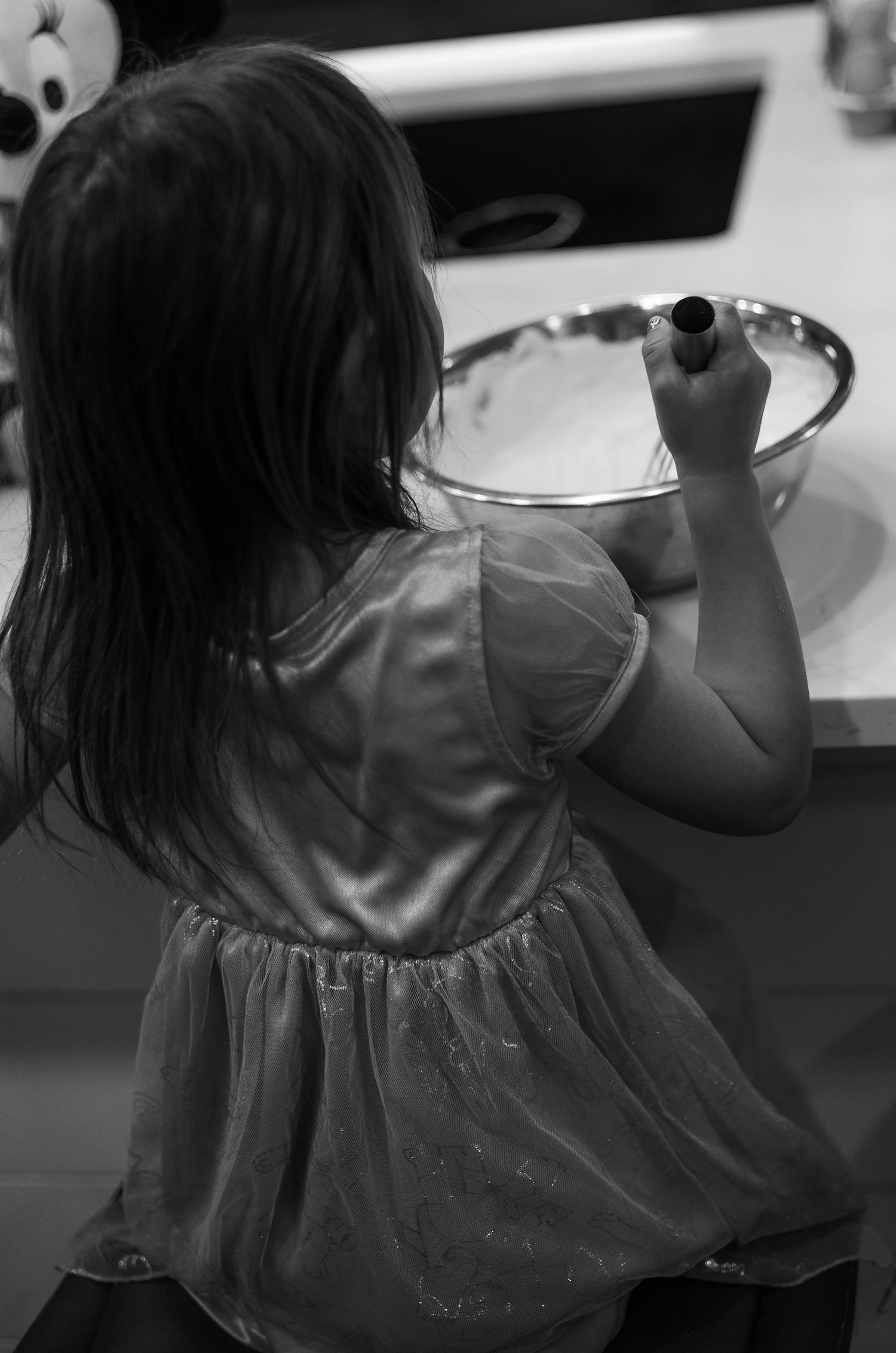 A child in a flowy dress is shown writing or drawing at a desk in black and white photography.