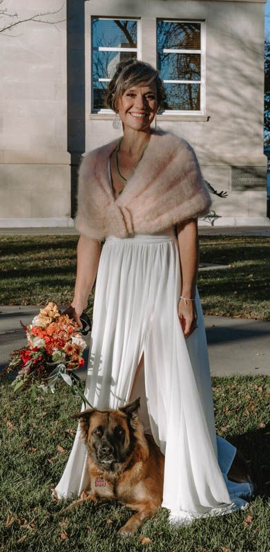 Person in white wedding dress and fur shawl poses with brown dog on grass.