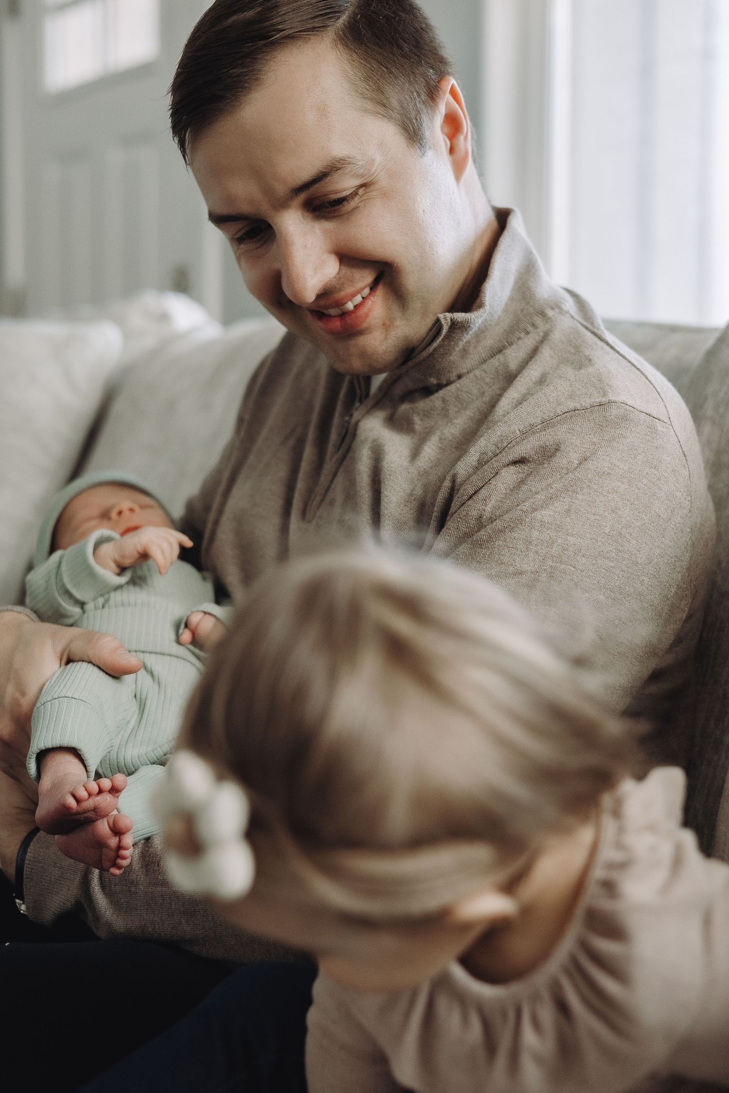 Parent in gray sweater holds and interacts with newborn baby and toddler in a softly lit room.