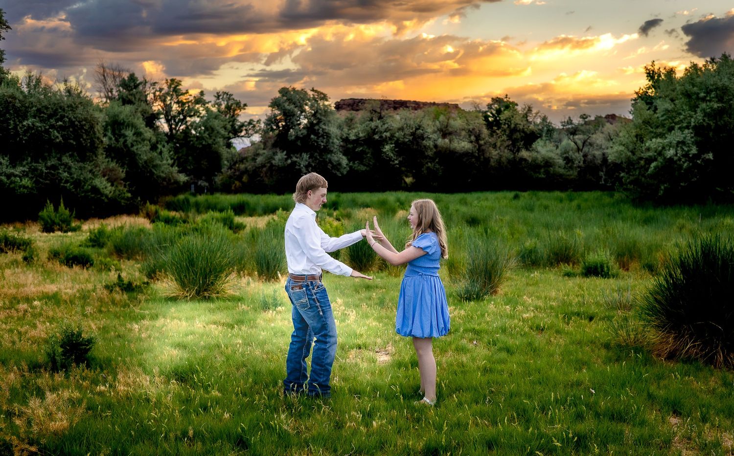 A couple dances joyfully in a lush green meadow at sunset, the man in jeans and the woman in a blue dress.