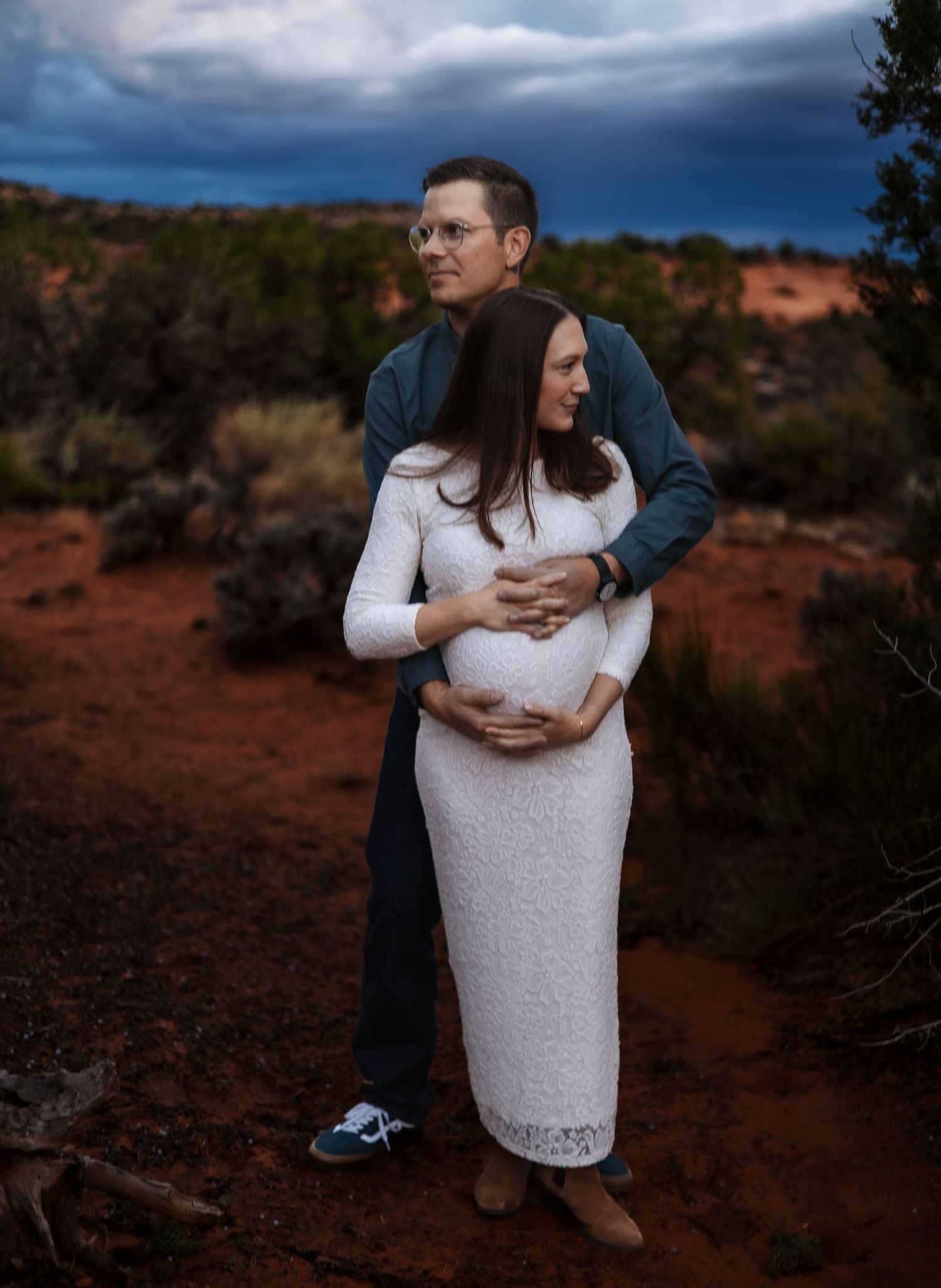 Couple embracing in red rock desert landscape, pregnant woman in white lace dress, dramatic stormy sky backdrop.