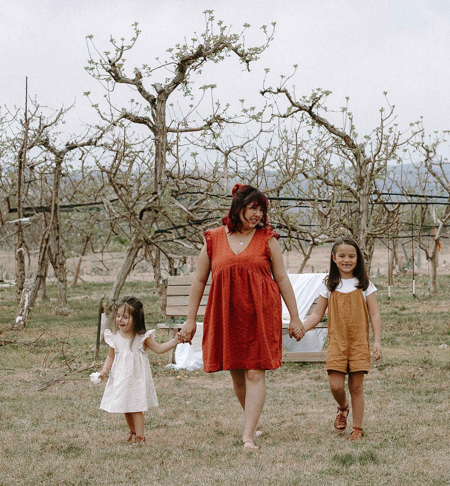 Children in rustic dresses walk and play among bare fruit trees in a pastoral orchard setting.