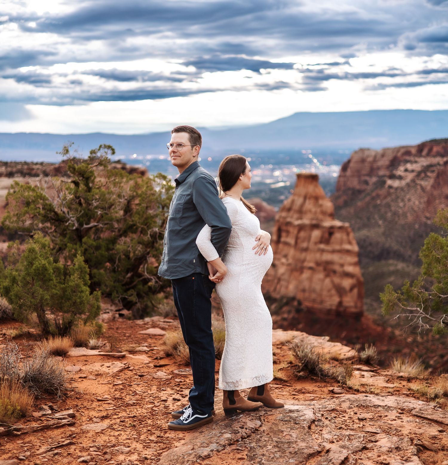 A couple sits on the edge of a red rock canyon overlook with dramatic cloudy skies above.