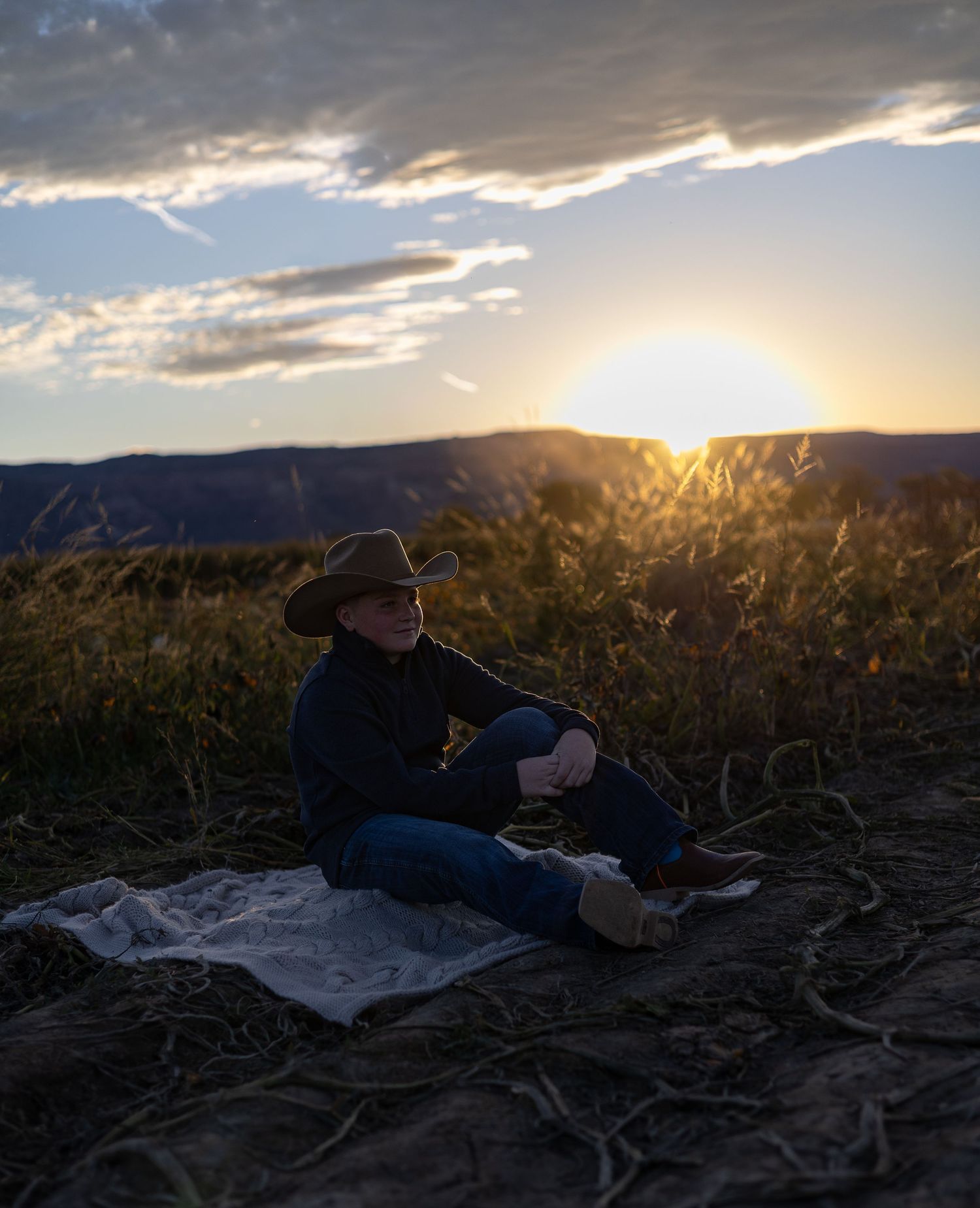 Person in cowboy hat sitting on blanket in field at golden sunset with mountains in background.