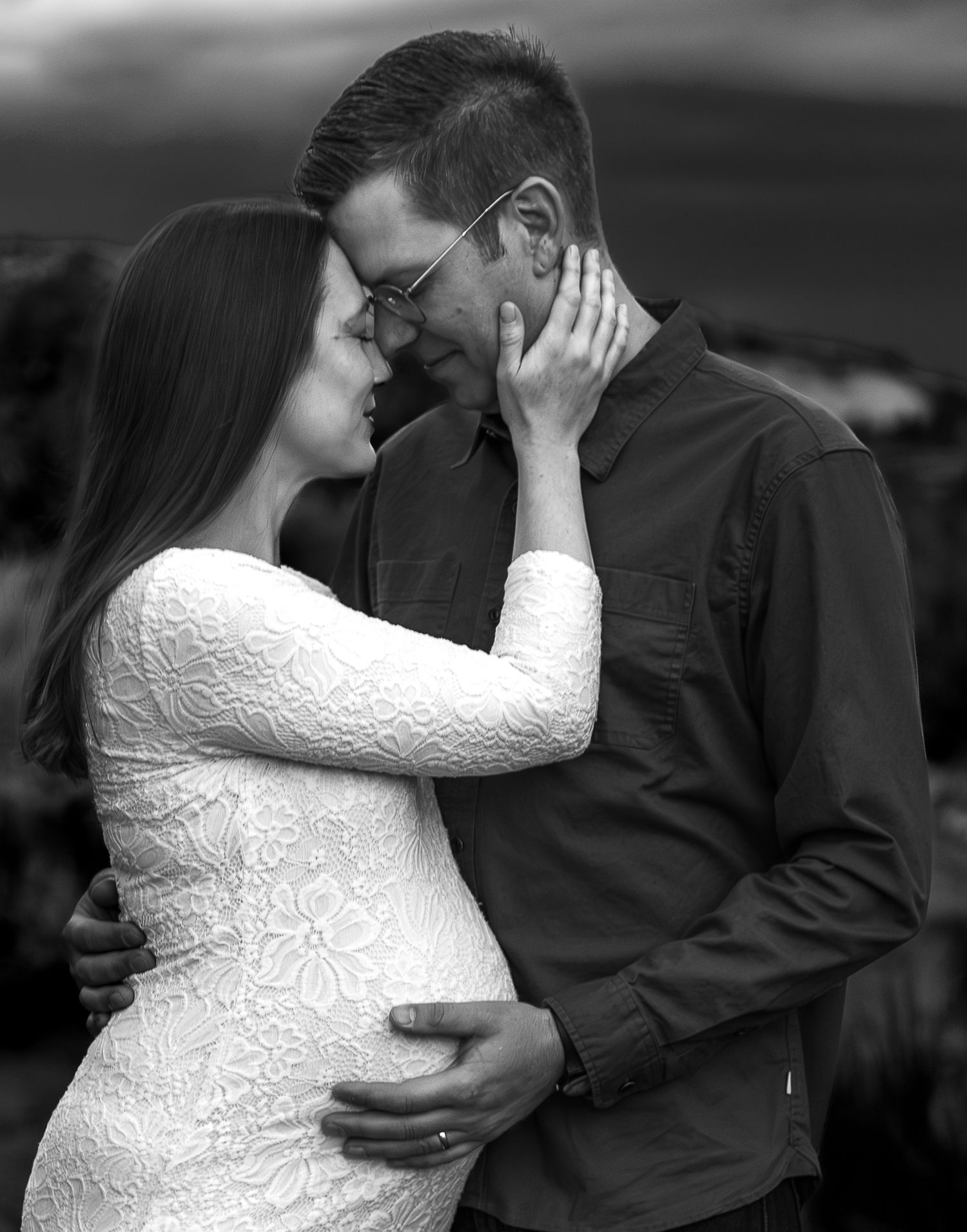 Black and white photo of a pregnant couple sharing a tender moment, woman in lace dress cradling bump.