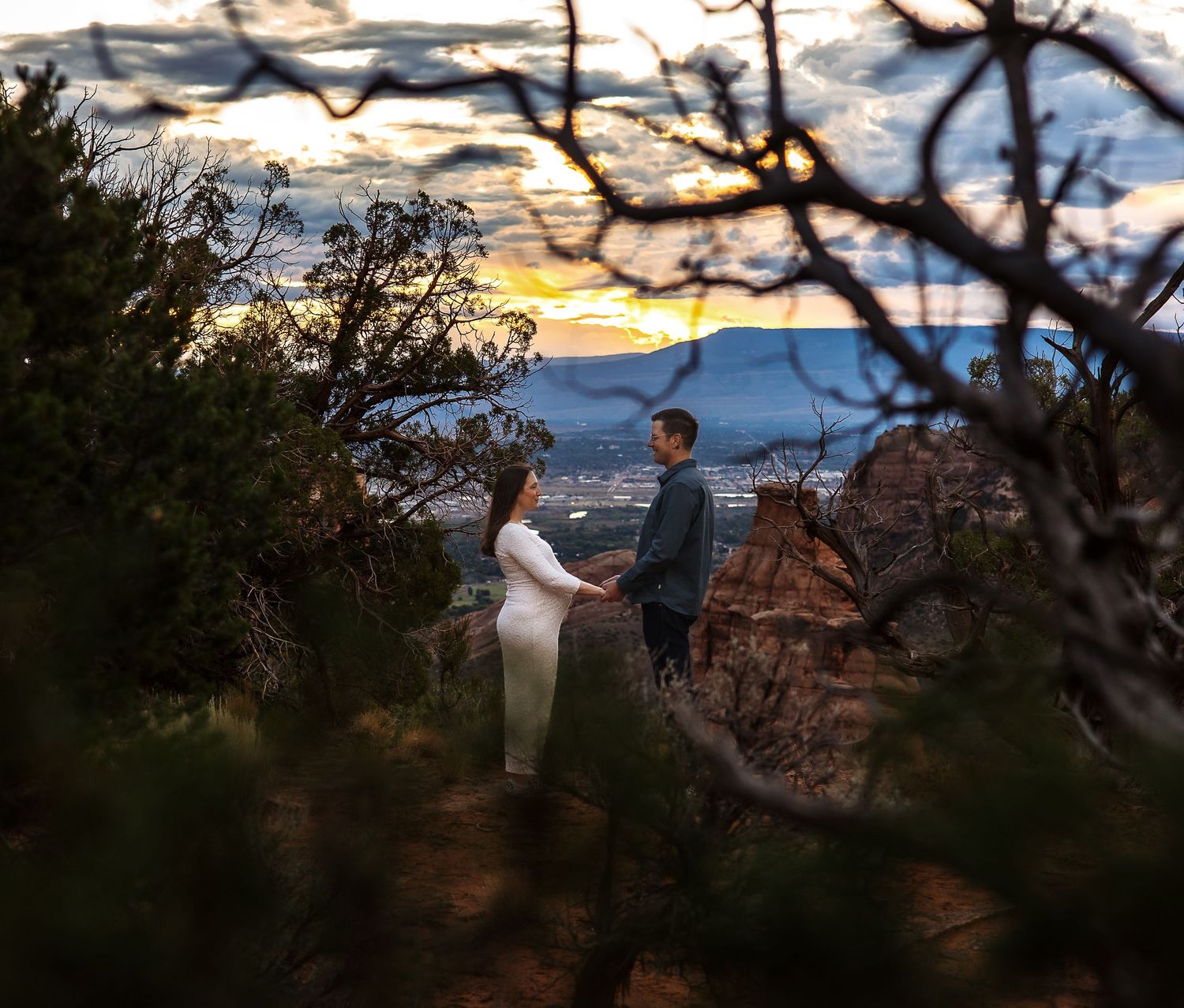 Silhouetted figures stand at the edge of a scenic canyon overlook during a dramatic sunset with orange and purple skies.
