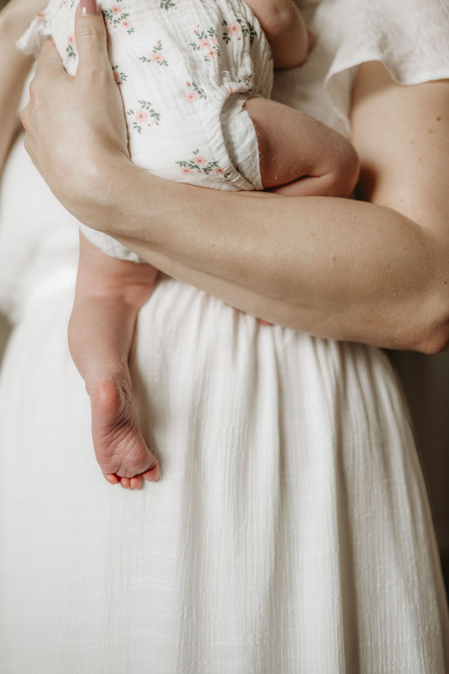 A mother in a white dress cradles a newborn baby in a floral outfit, with tiny feet visible.