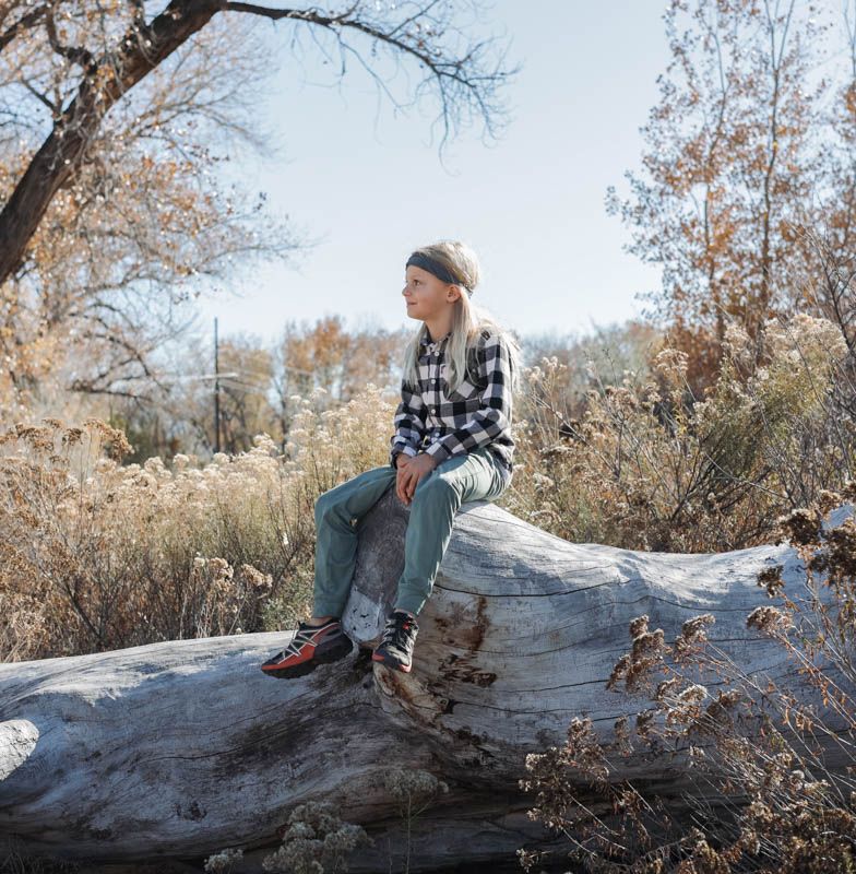 Someone in a plaid shirt and beanie sits thoughtfully on a fallen log during autumn in a natural outdoor setting.