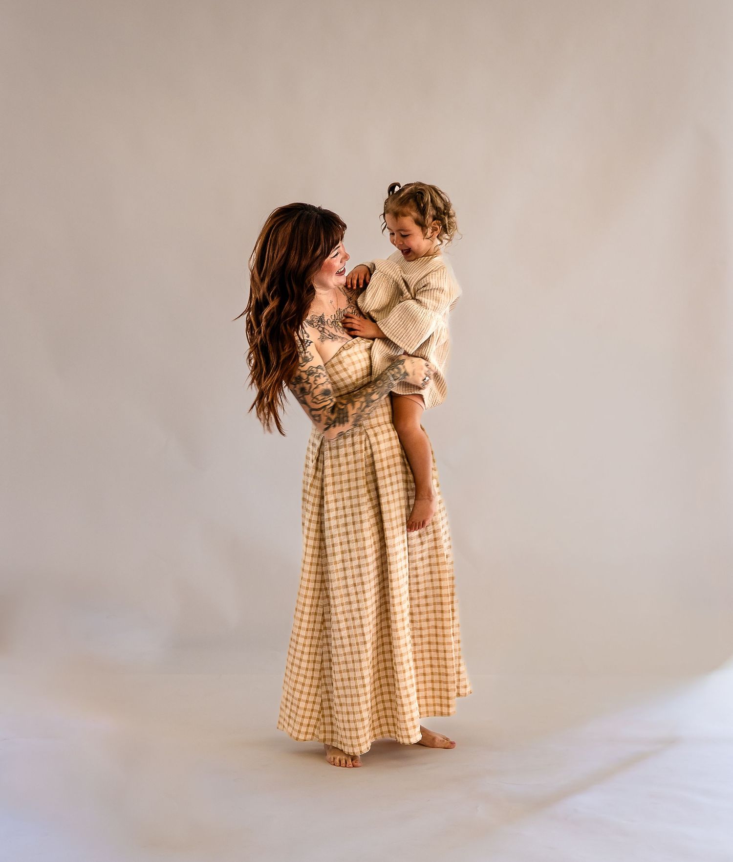 A woman in a gingham maxi dress holds a toddler in a studio setting, both smiling warmly.