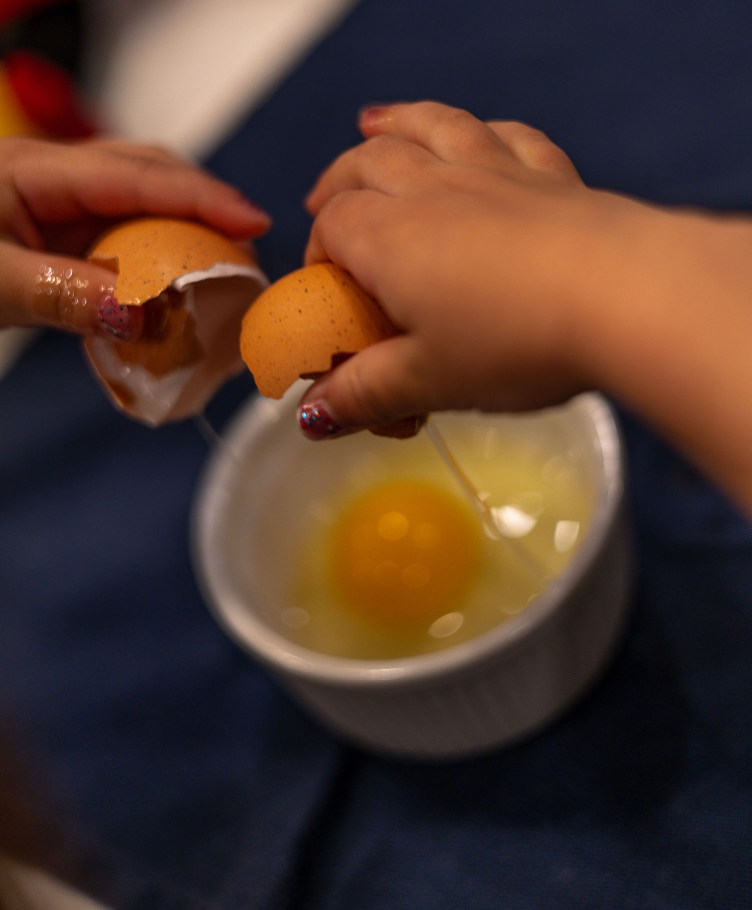 Hands cracking an orange egg into a white ceramic bowl against a dark background.