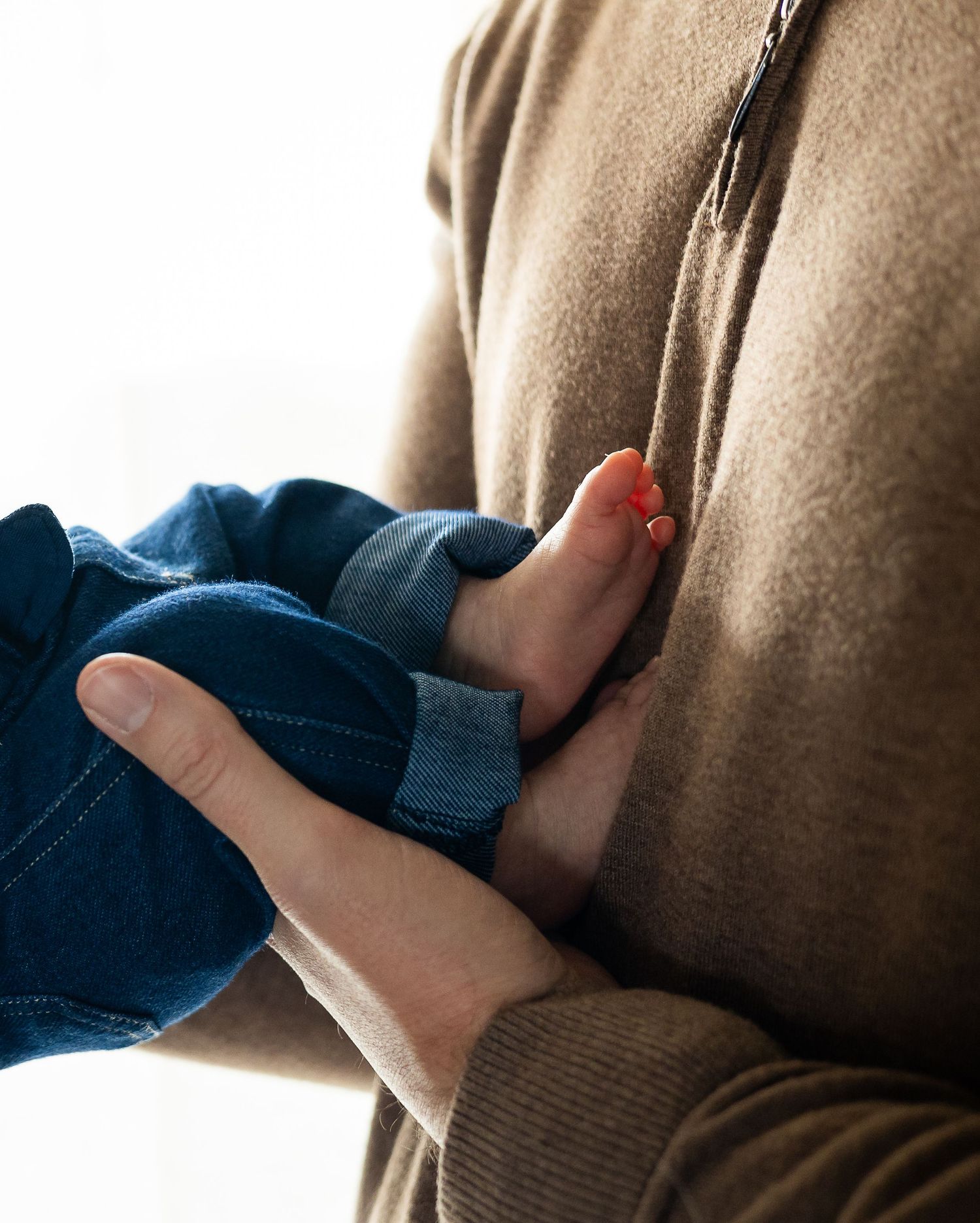 A tender moment shows a person holding and kissing a small baby while wearing a beige sweater in a bright setting.