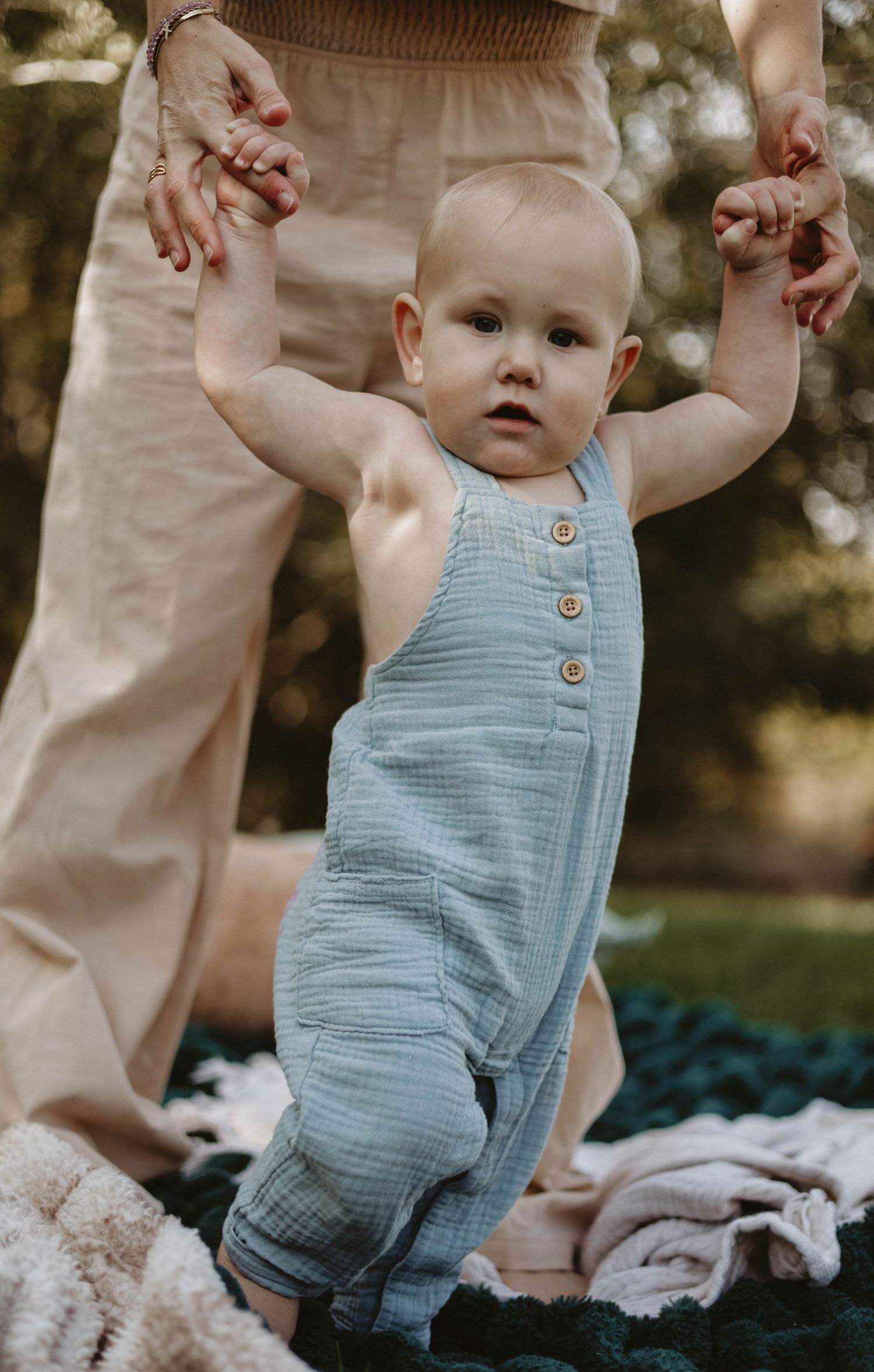 Baby wearing blue romper supported while standing on blanket outdoors.