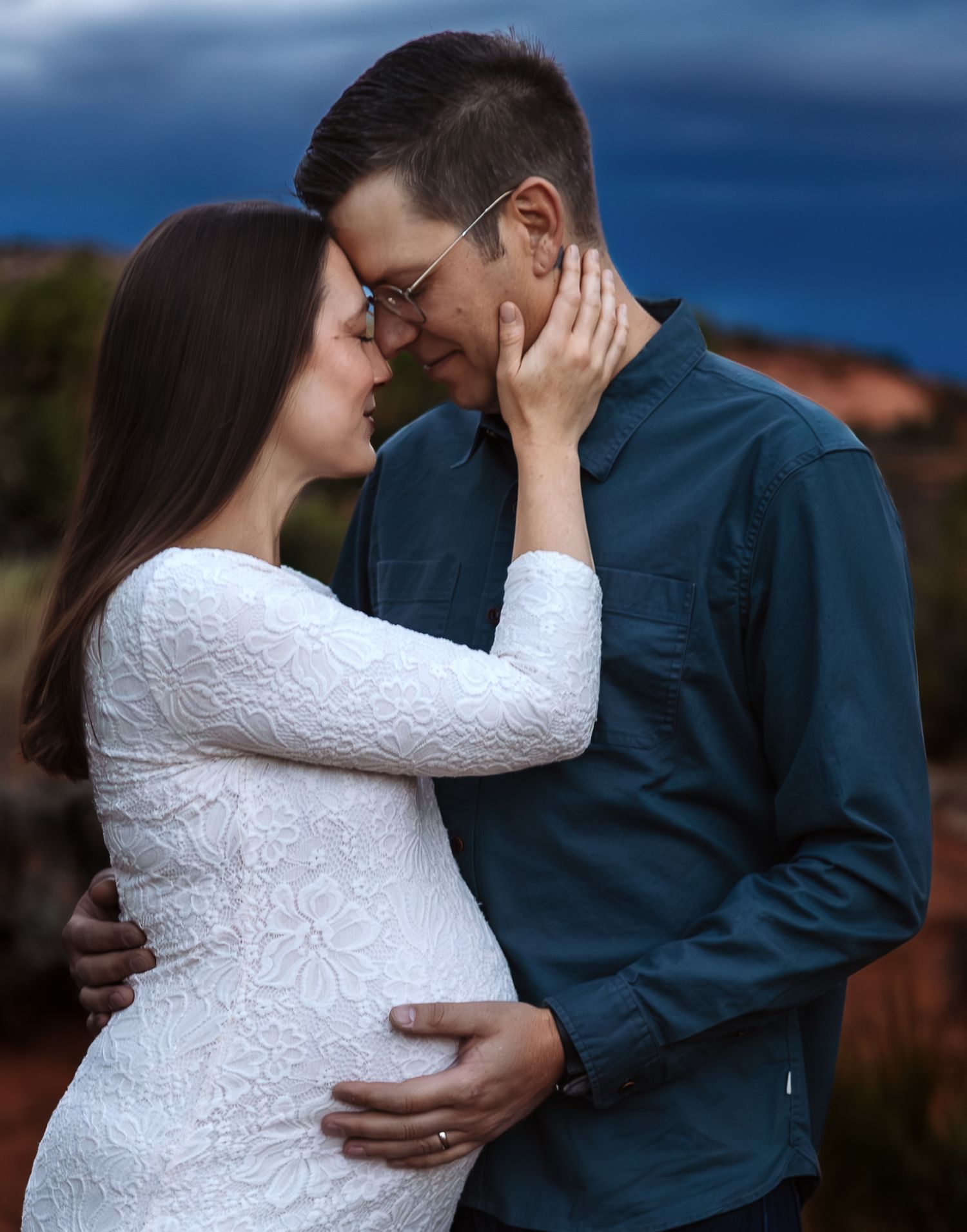 A romantic couple in formal attire share an intimate embrace against a blue evening sky background.