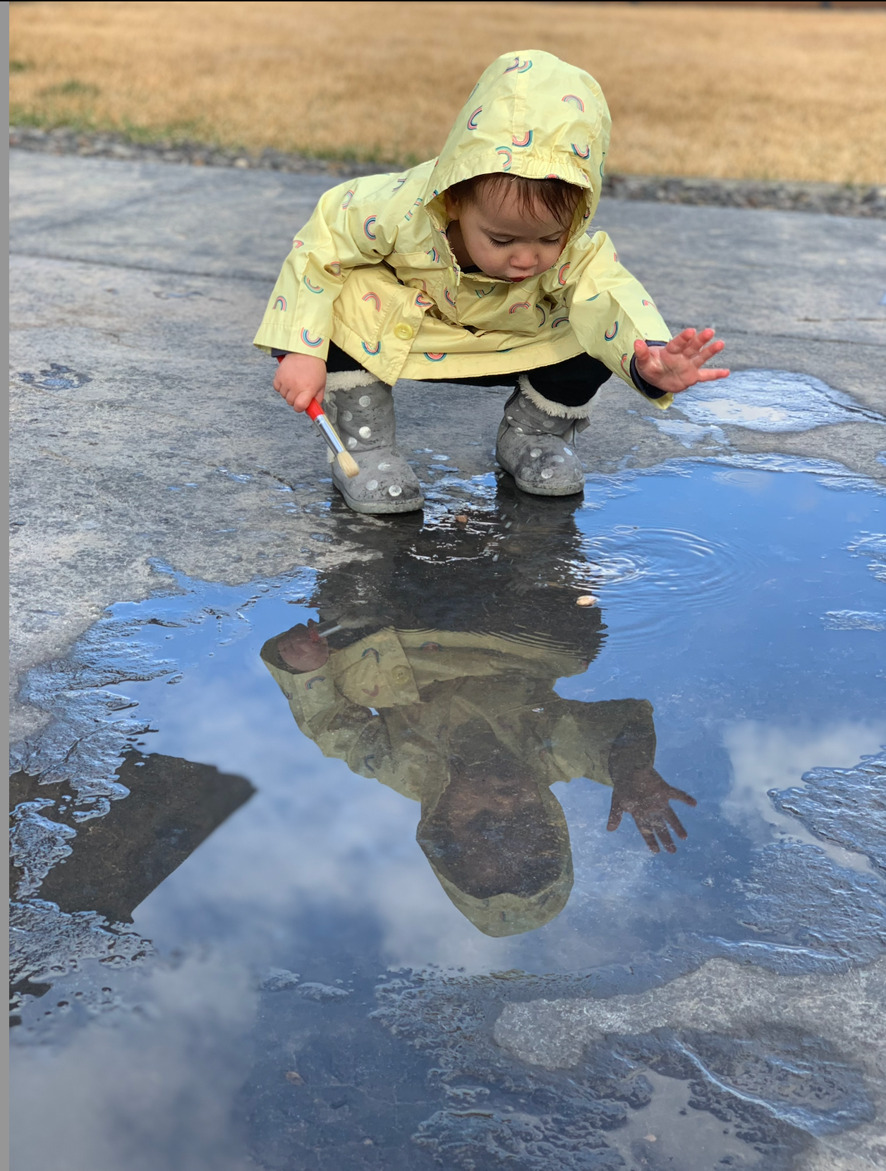 Someone in a yellow raincoat crouches by a puddle watching their reflection in the water on a rainy day.