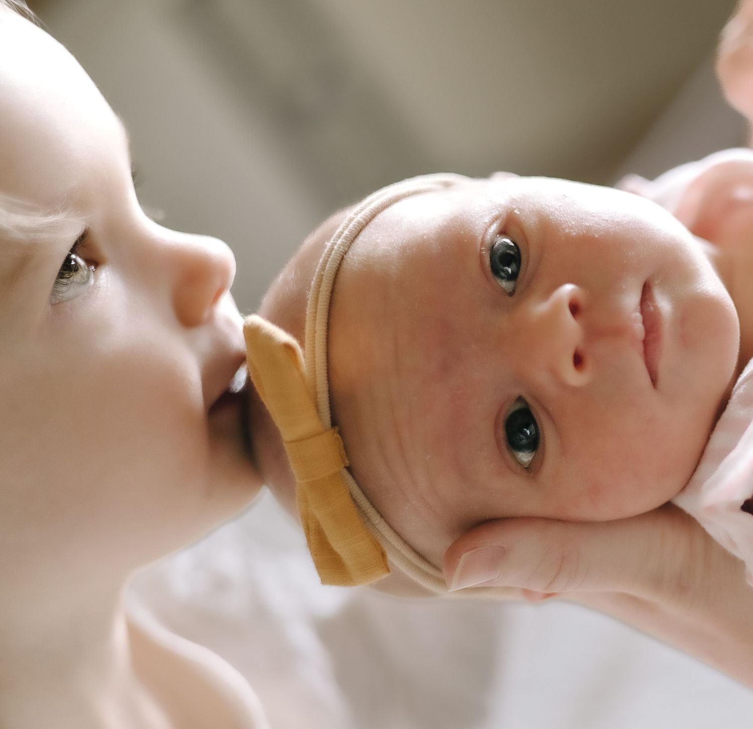 A baby lying on white bedding looks up with bright eyes while wearing a tan headband.