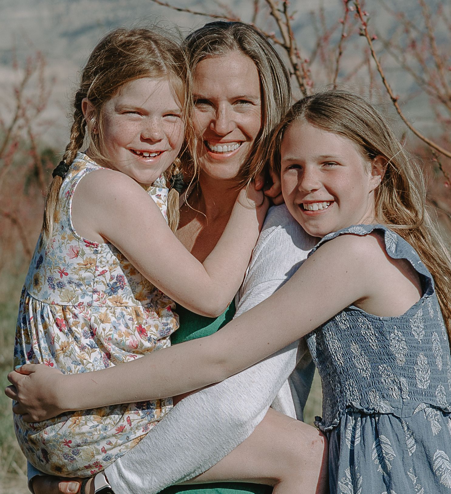 A loving embrace between siblings in coordinating gray and floral dresses is captured in a sweet outdoor moment.