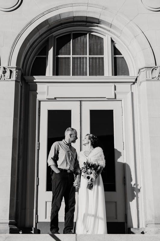 A couple in wedding attire stand together under an elegant arched doorway in black and white.