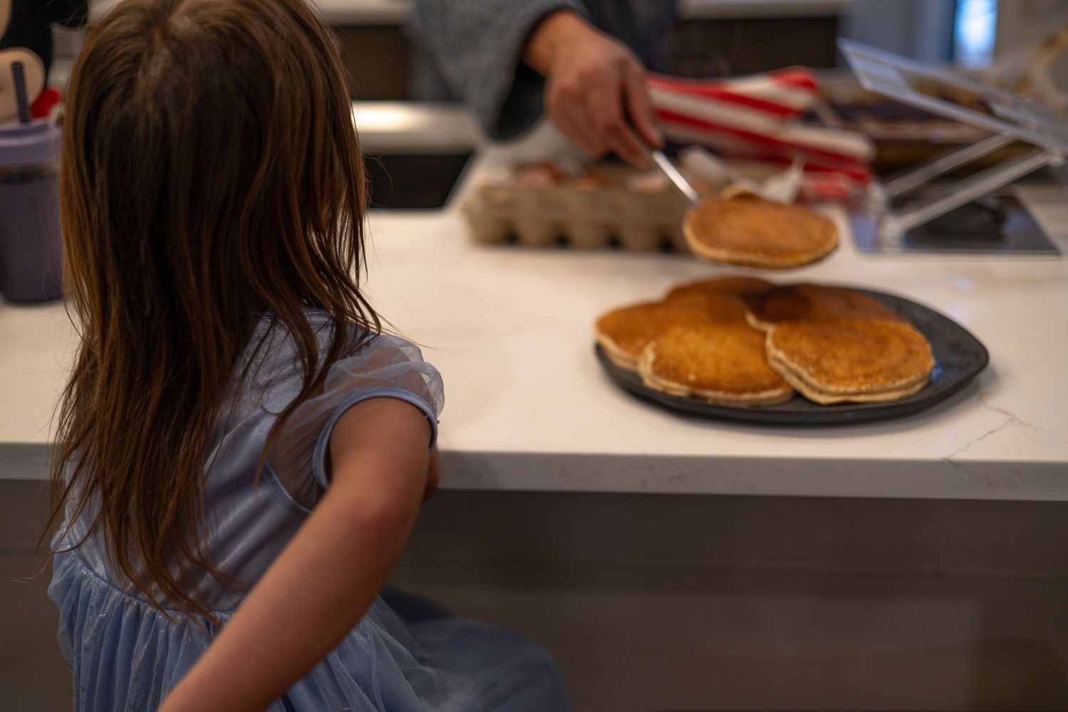 Fresh golden pancakes on a dark plate with someone in a blue dress sitting at a kitchen counter.