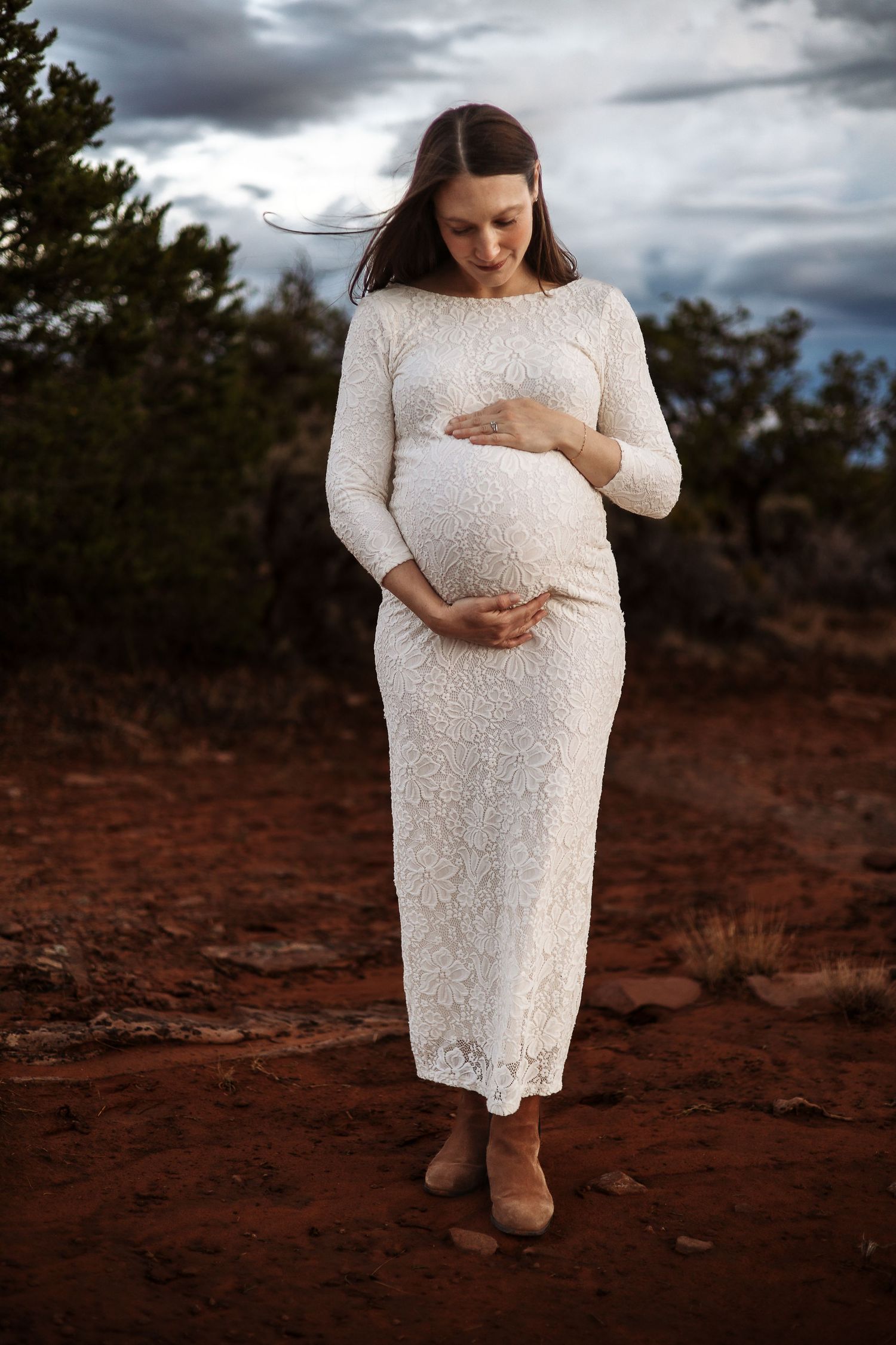 A maternity photoshoot in a white lace dress against a dramatic cloudy sky and red desert landscape.
