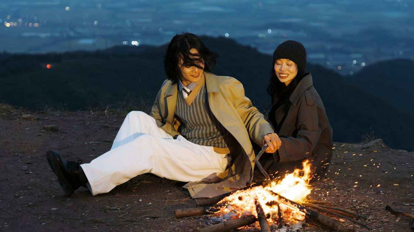 A romantic couple sitting by a campfire on a hilltop at dusk overlooking a glittering city skyline.