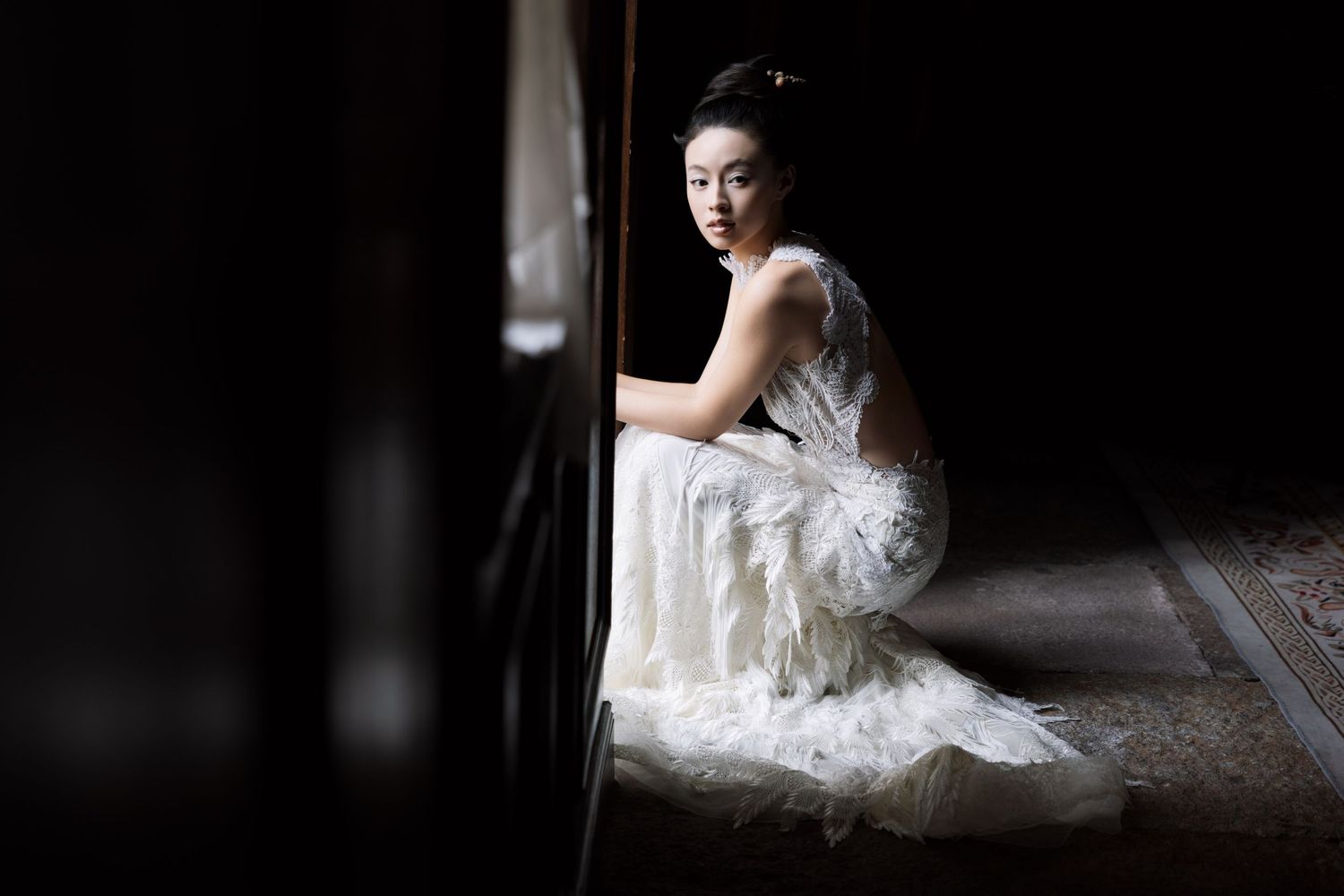 A woman in an elegant white feathered gown crouches beside a dark wall, gazing at the camera in a moody, dramatic portrait.