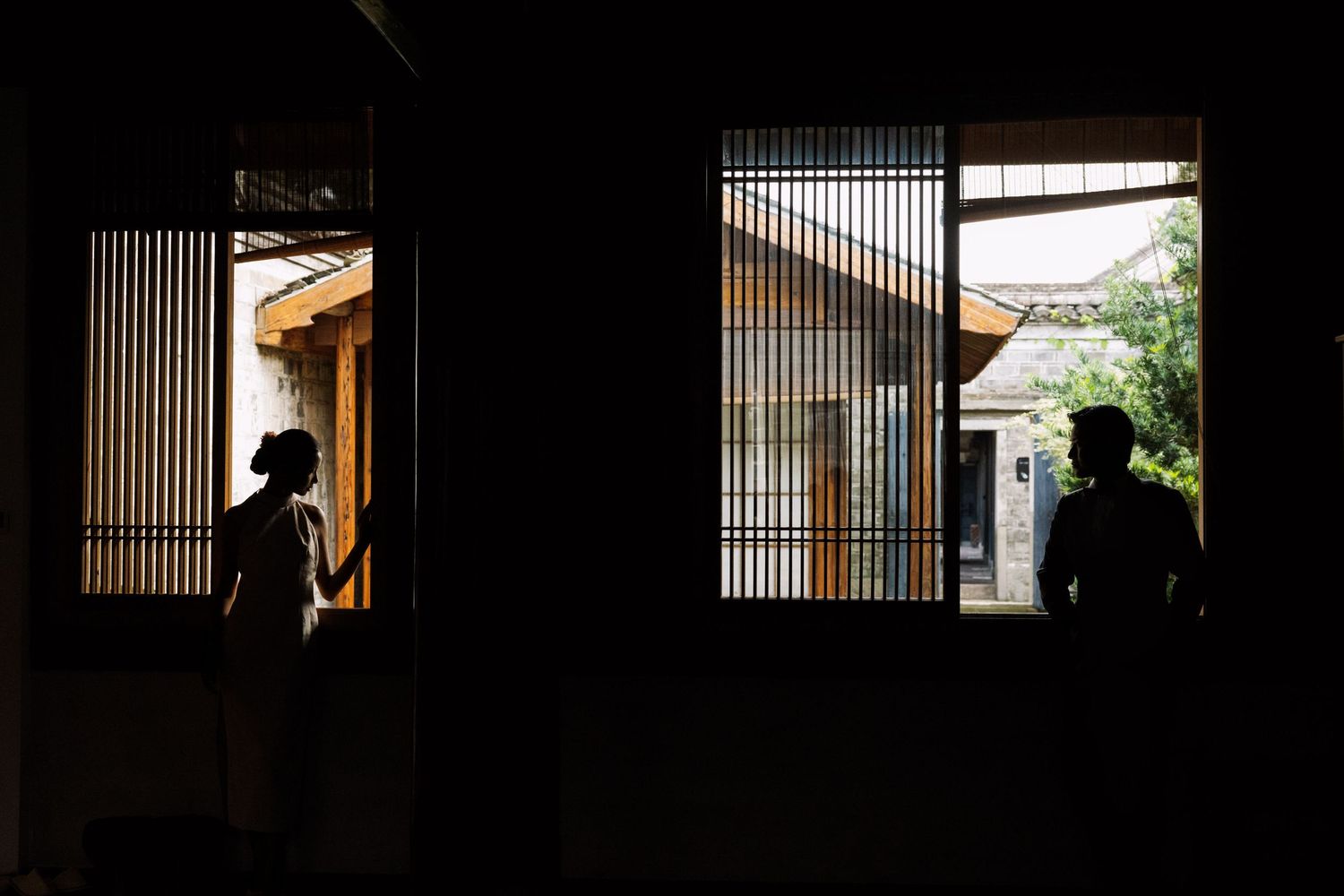 Two silhouetted figures stand in a dark room beside bright lattice windows overlooking a traditional wooden building and green trees.
